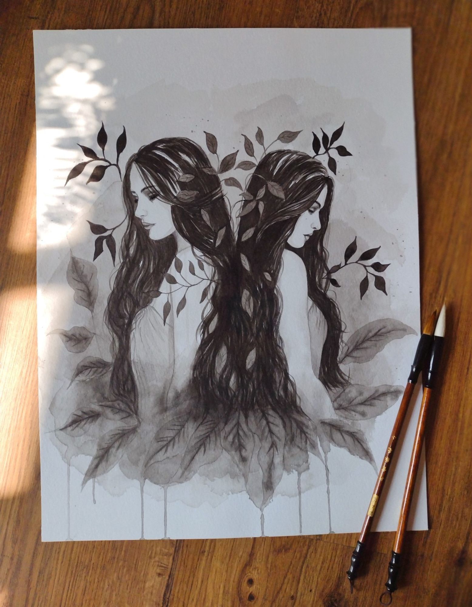 Ink wash drawing of two women standing back to back and surrounded by leaves. The drawing is laying on the floor with 2 brushes on the side.