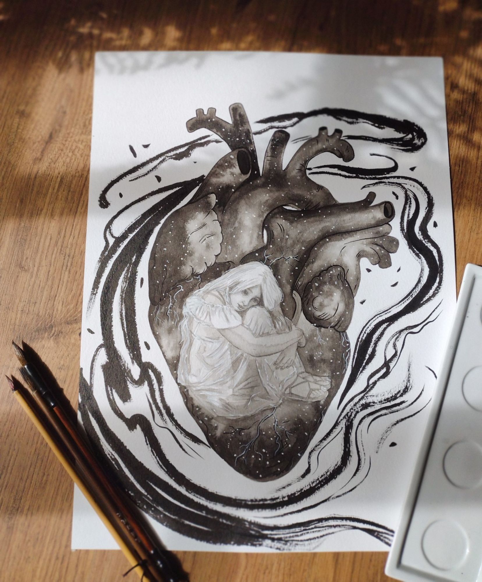 Ink wash drawing of a girl sitting inside of an anatomical heart, holding her knees. The heart is painted as a night sky with stars. The drawing is laying on the floor with paint brushes on the left side of it.