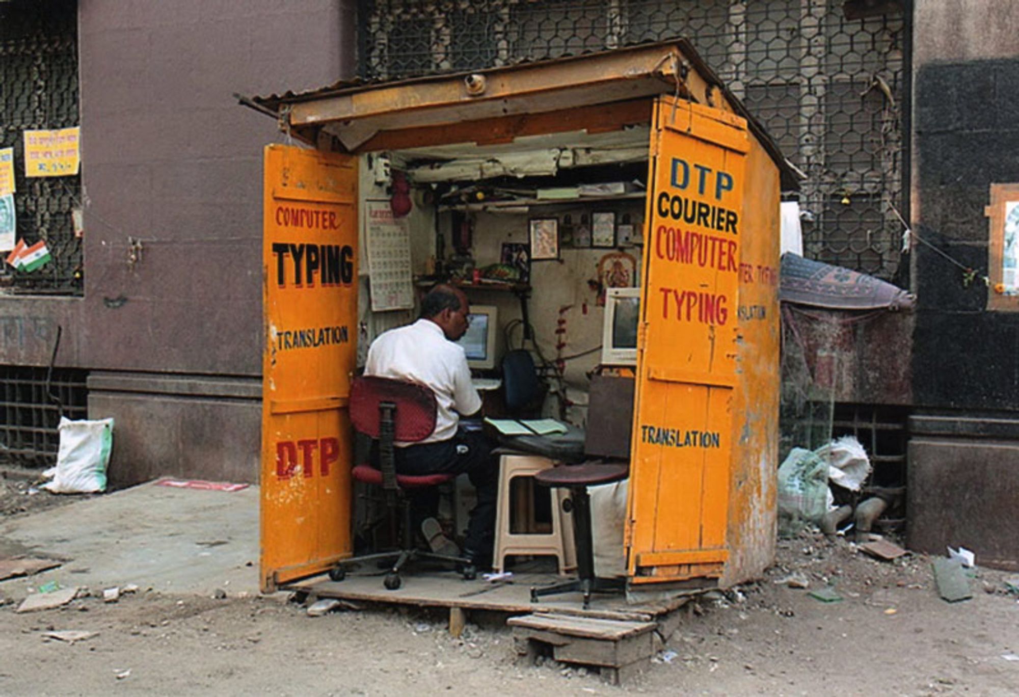 A man sitting in a dingy yellow shack filled with computer equipment and misc computer things. He is looking at some paperwork next to him typing on his main computer. The sides of the yellow shack have "DTP courier computer typing translation" and "computer typing translation DTP" on the sides. The shack appears to be situated in a dirty alley way or on the side walk of some street against the side of a building.