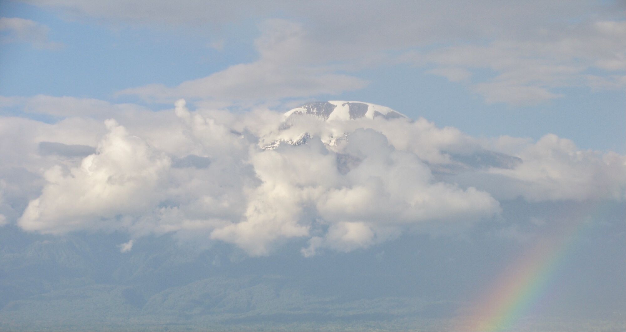 Schneebedeckter Berg schaut aus den Wolken, rechts unten ein Regenbogen