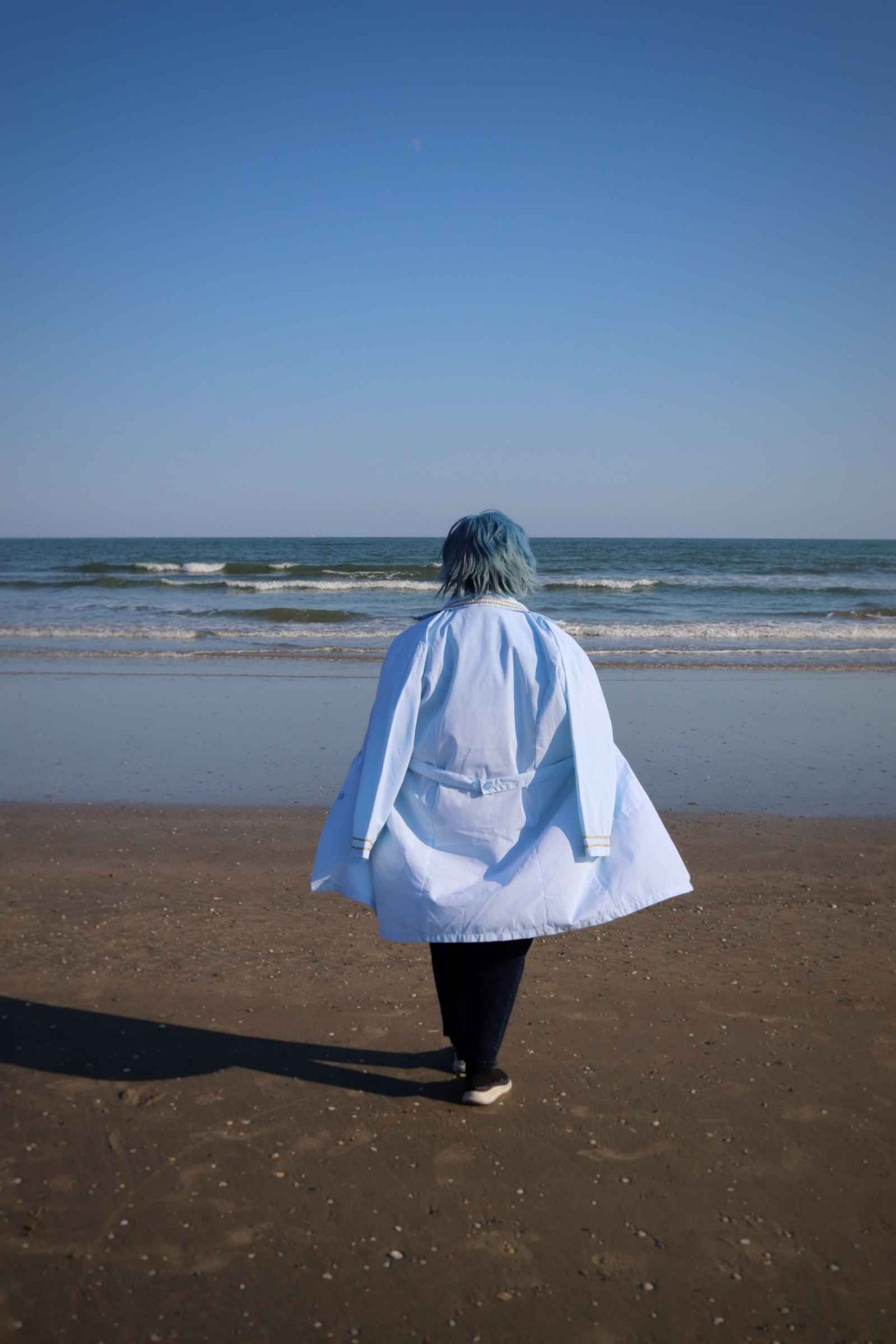 A cosplayer dressed as a doctor walks in the middle distance on a flat beach toward the ocean.