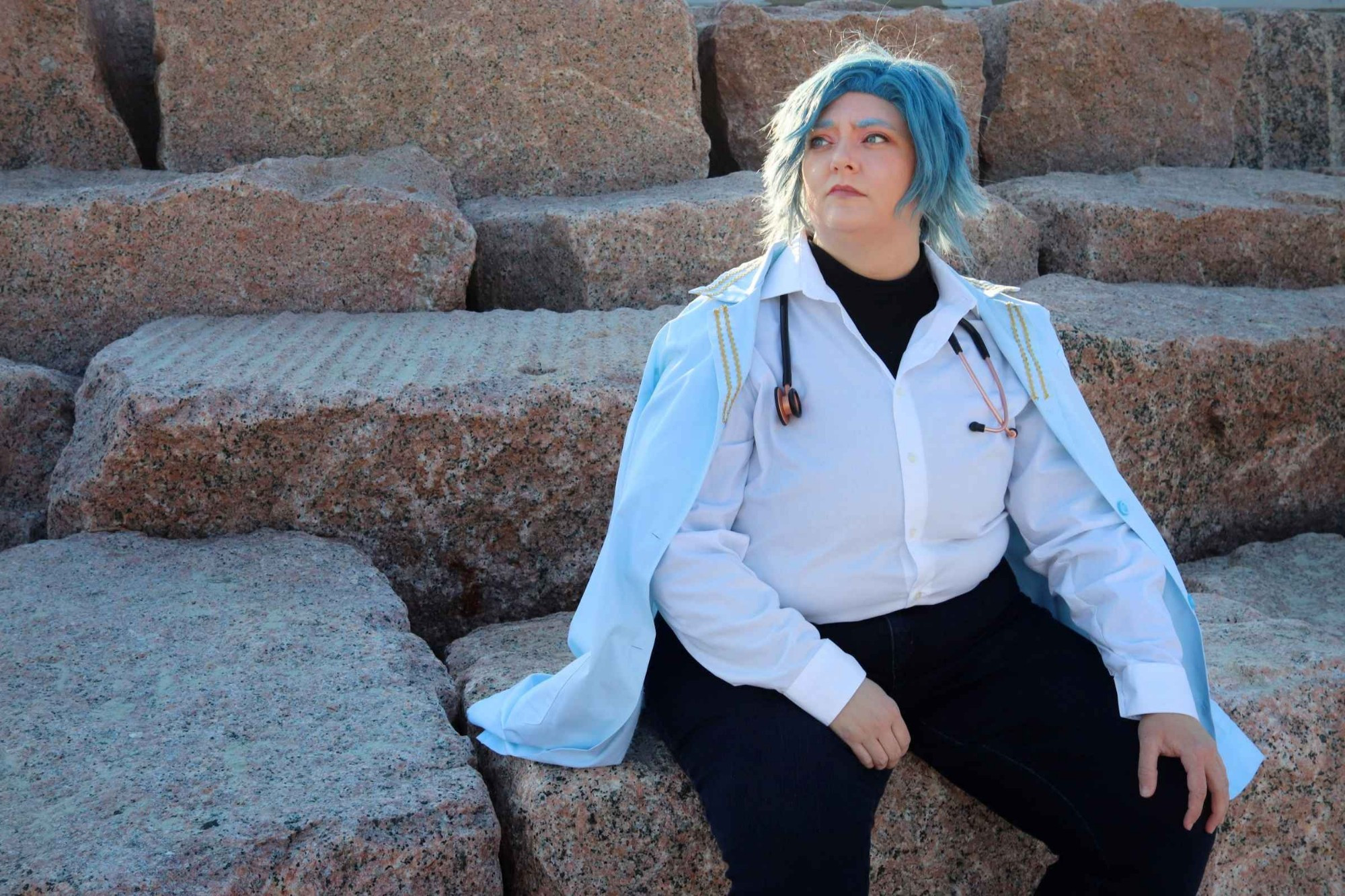 Cosplayer sitting on sandy granite steps in a blue wig, white button up shirt, dark blue pants, and light blue lab coat. Seems to be a doctor!