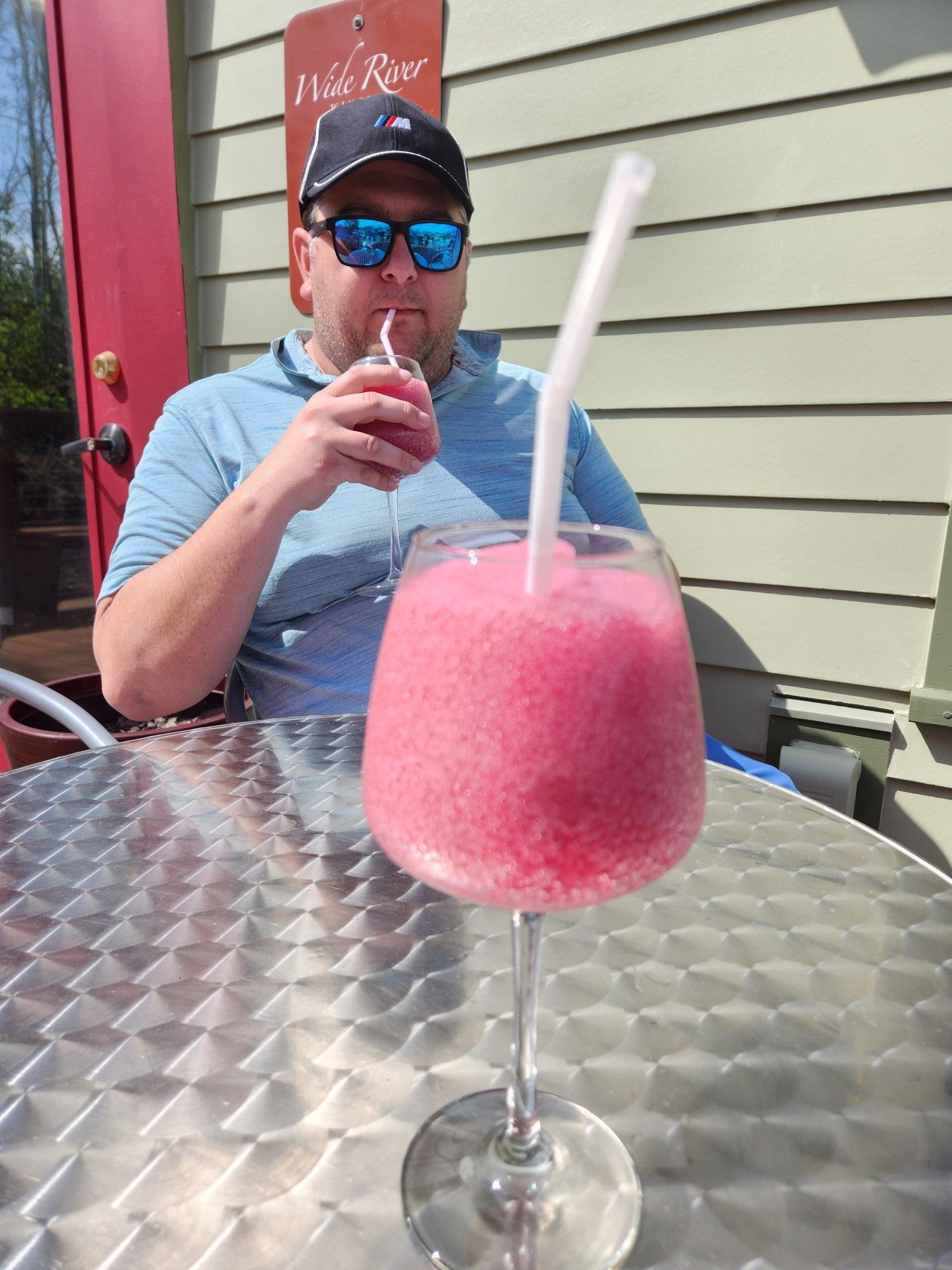 A wine glass full of pink wine slushy sitting on a silver reflective table. A white dude wearing a light blue golf polo, an M3 hat, and polarized sunglasses is sitting across the table sipping his own wine slushy.