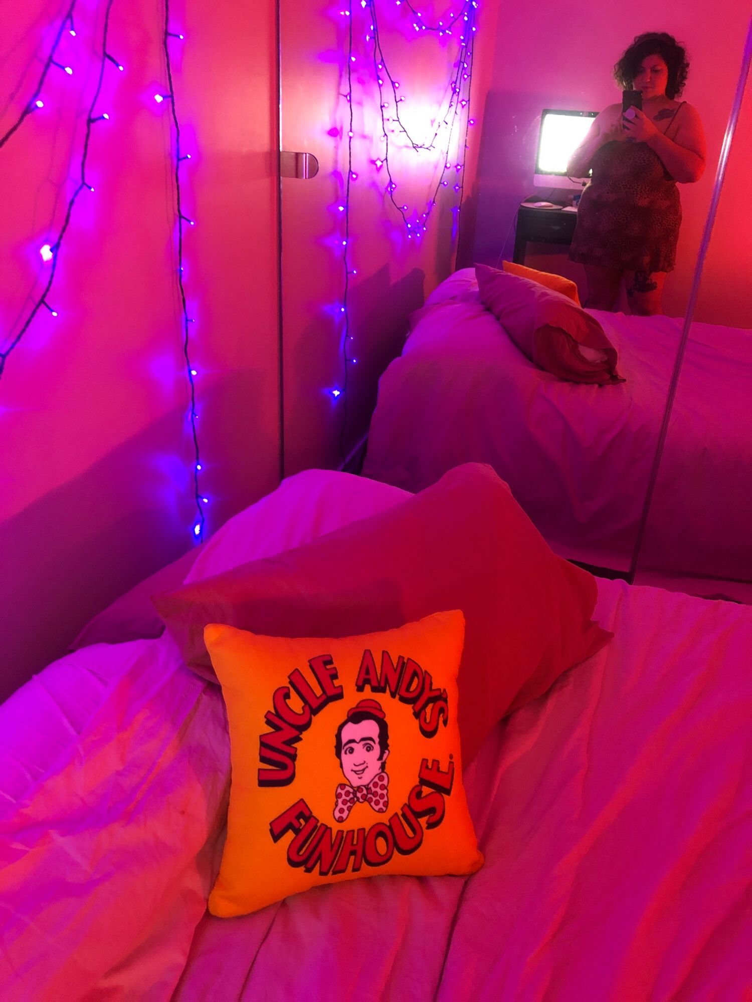 Pic of me in my bedroom mirror reflection, UV fairy lights on the wall and bathed in the light of a pink neon sign (not pictured). On my bed is a cushion with Uncle Andy’s Funhouse logo on it (Andy Kaufman)