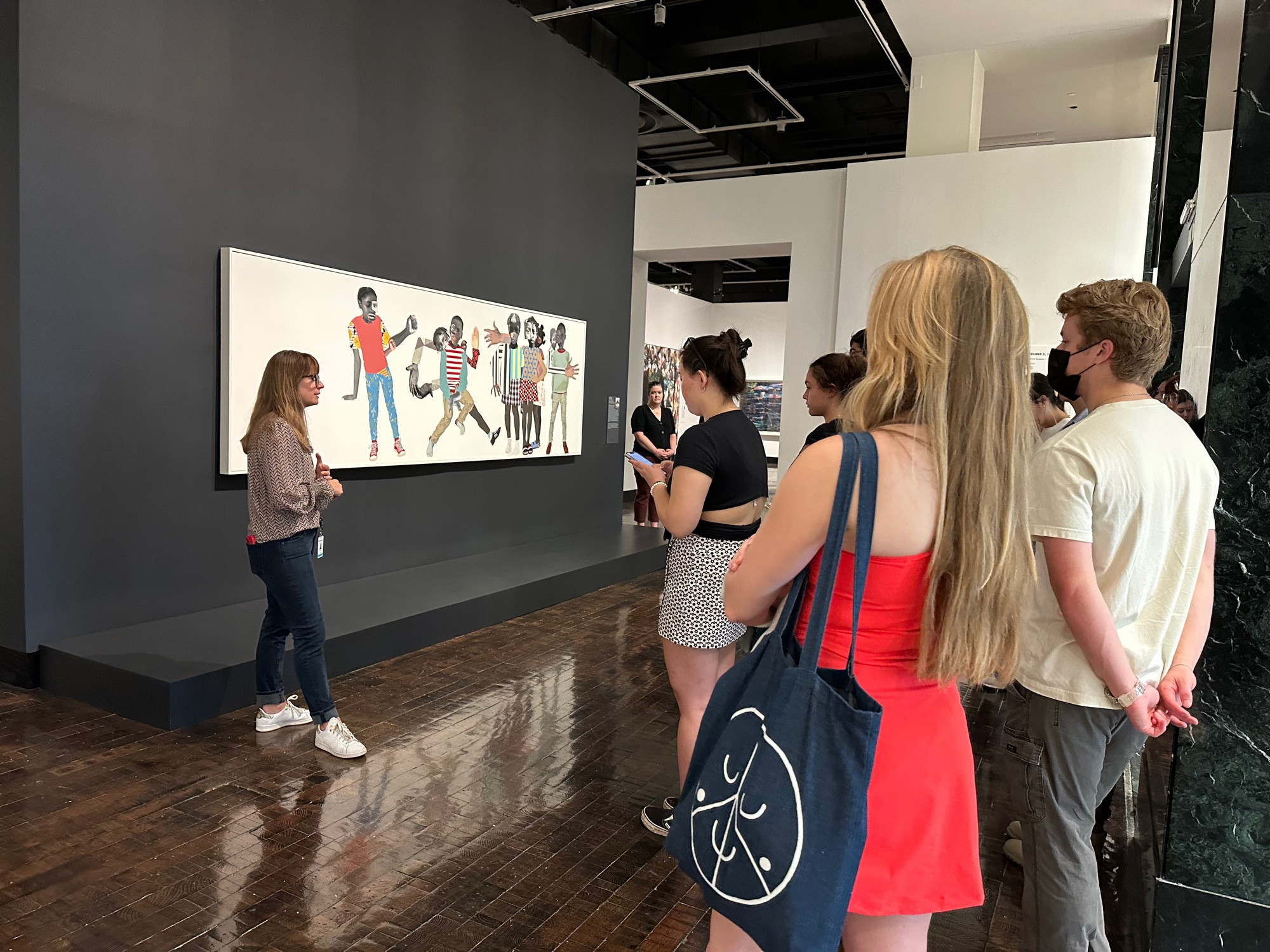 A photo of an interior exhibition space showing a group of college students on the right and a woman with long golden hair and glasses on the left. The woman on the left is speaking to the group of students and stands in front of a large work of art in canvas form on the wall.