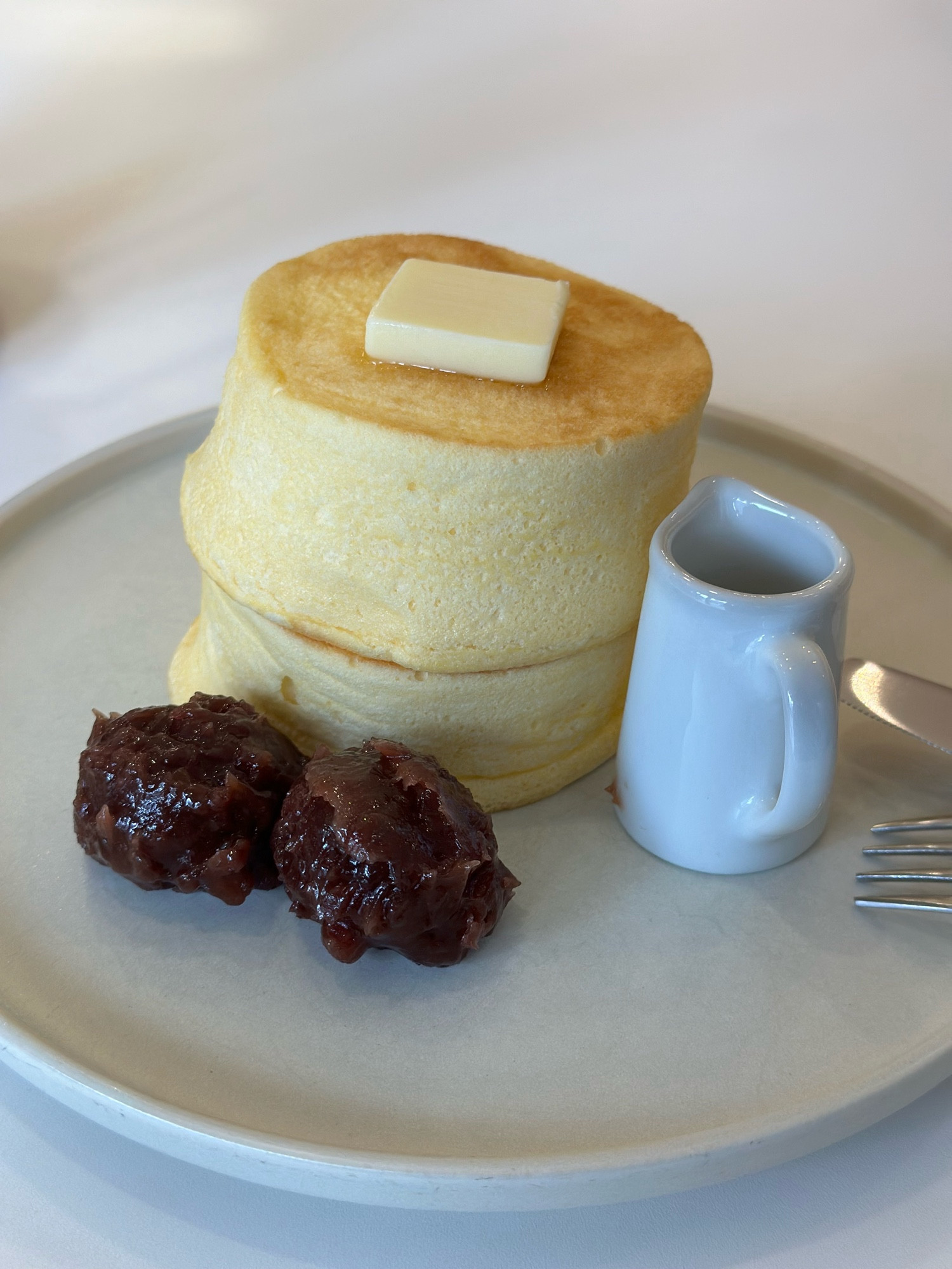 Two golden-topped fluffy pancakes with a pat of butter on top of them on a plate with two scoops of red bean paste and a small container of maple syrup next to them.