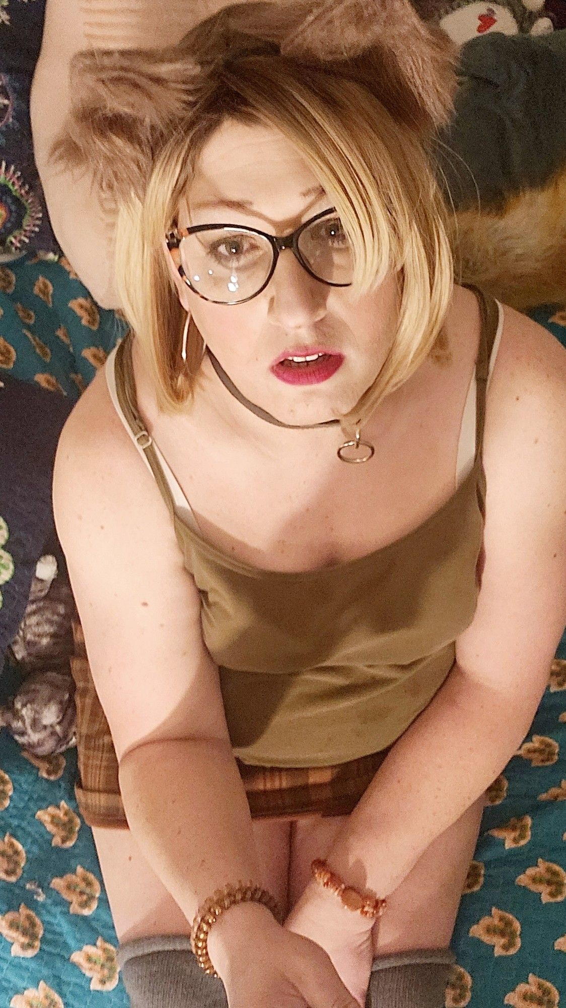 overhead angle pic of me looking up at you pleadingly, blonde with glasses and tan cat ears, red lipstick, black and chrome kink collar, hunter green strappy tank top, brown plaid skirt, grey thigh high socks, sitting on my bed