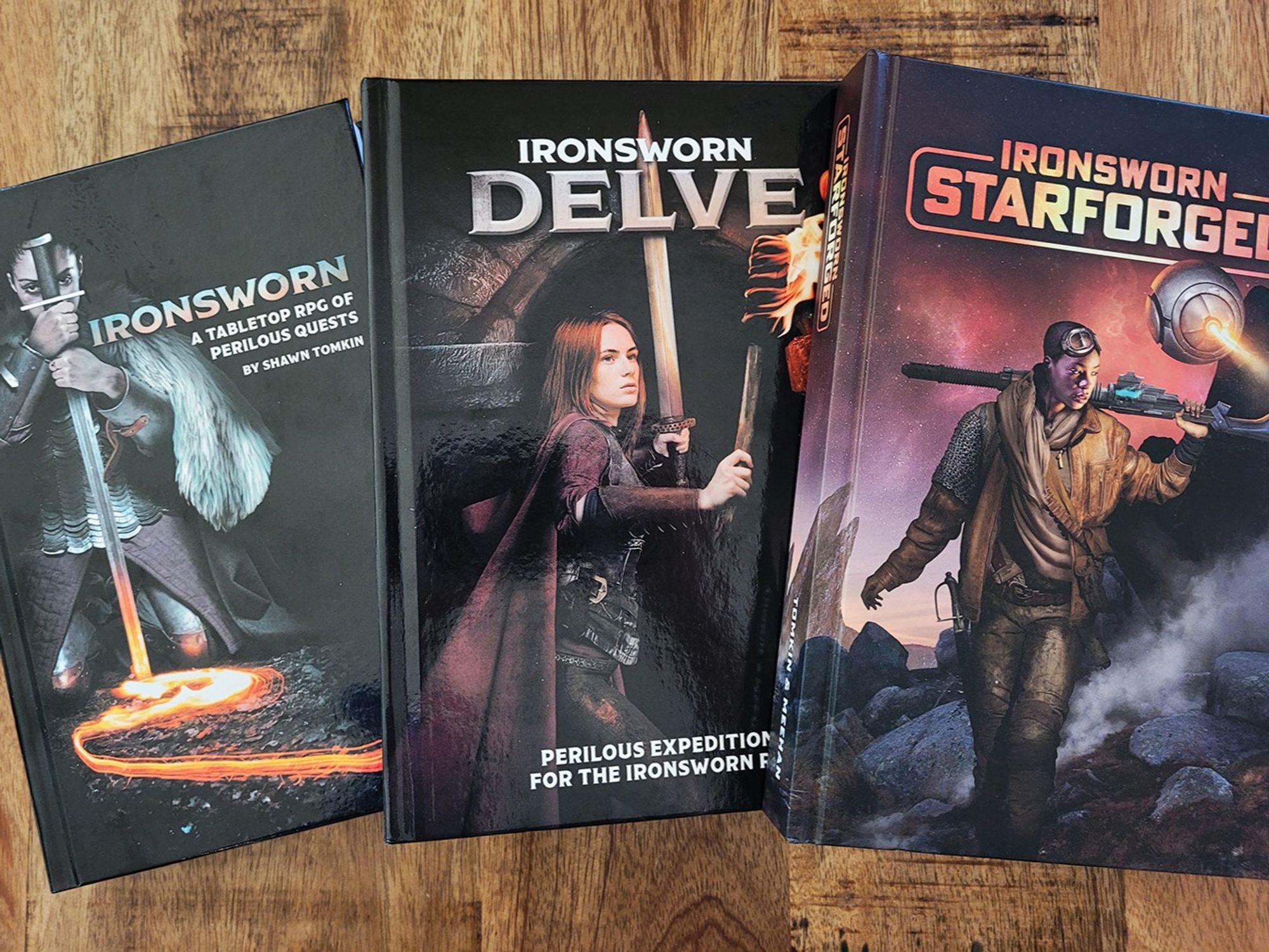 A photo of a set of books sitting on a wooden table. The books are titled Ironsworn, Ironsworn: Delve, and Ironsworn: Starforged. 