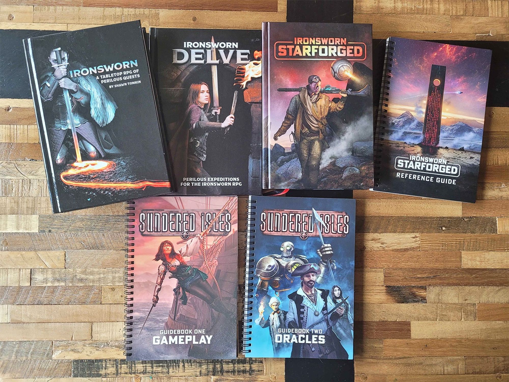 A photo of an array of books on a wooden table. The books include Ironswsorn, a Tabletop RPG of Perilous Quests, Ironsworn: Delve, Ironsworn: Starforged, the Ironsworn: Starforged Reference Guide, and Sundered Isles Guidebook One and Two.