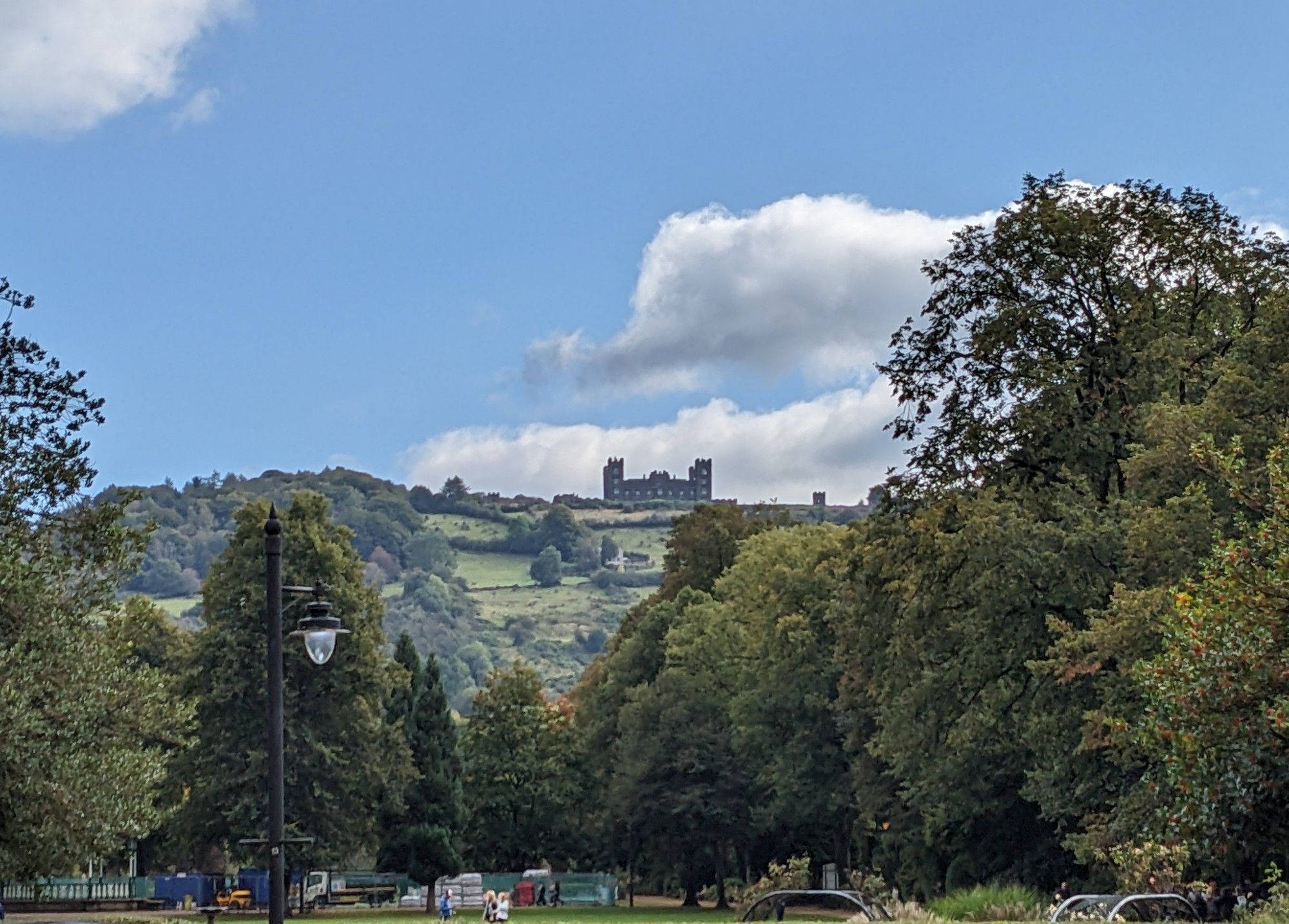 Photo of Riber Castle taken from central Matlock. The castle is at the top of a wooded hill. Blue skies and a couple of white clouds