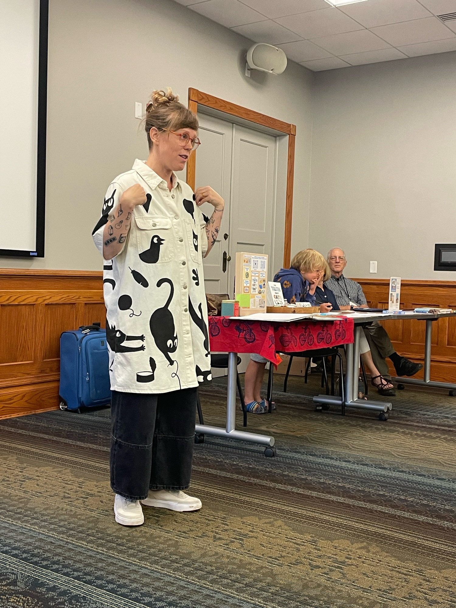 Lucy Knisley speaking to an audience. She is wearing a thrifted white coat covered in black cat illustrations of her own design