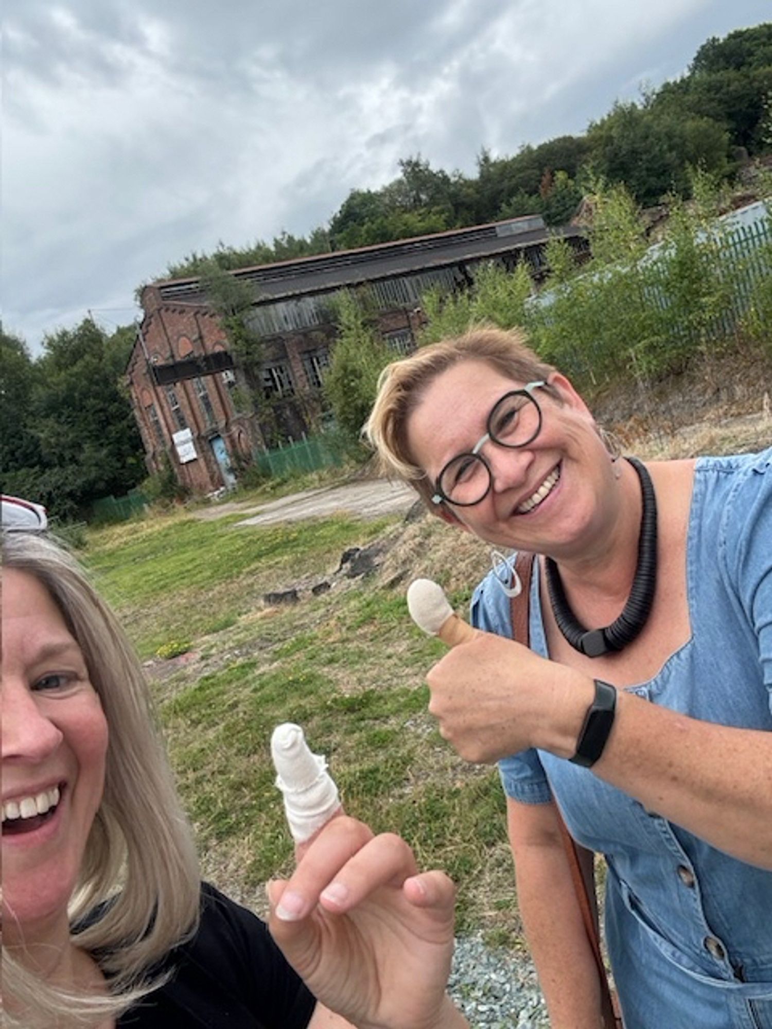 Two laughing women one with a bandaged thumb and the other with bandaged finger with large derelict building in background