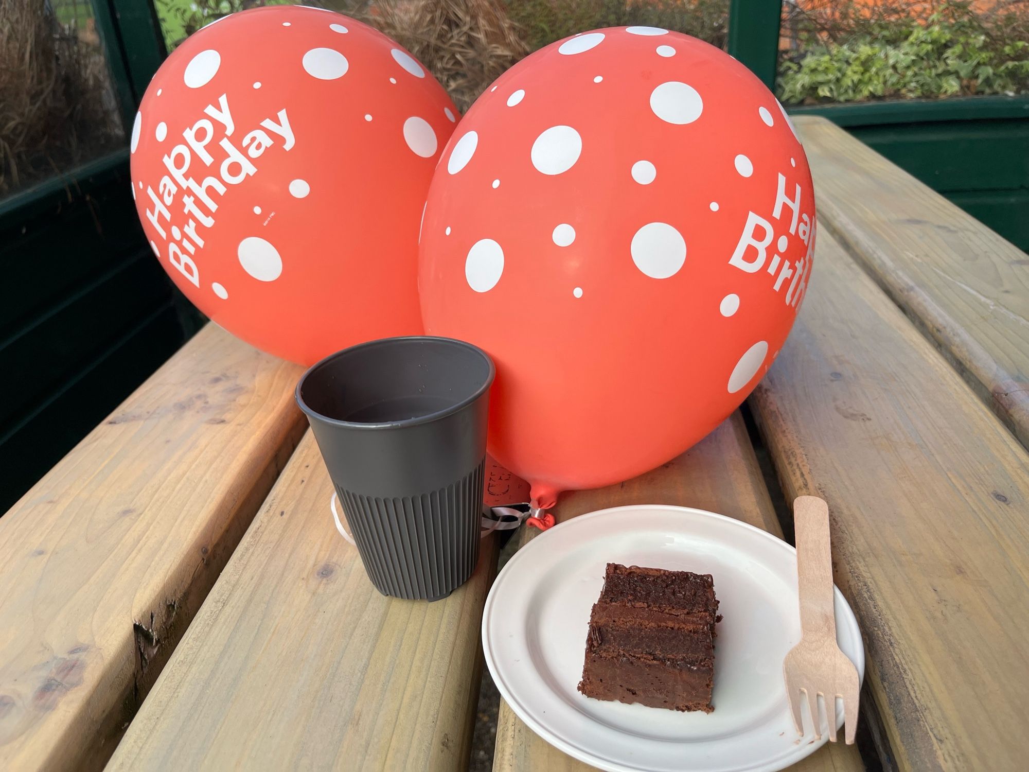 Two red ‘happy birthday’ balloons, tea & a slice of chocolate cake on a wooden table after Parkrun