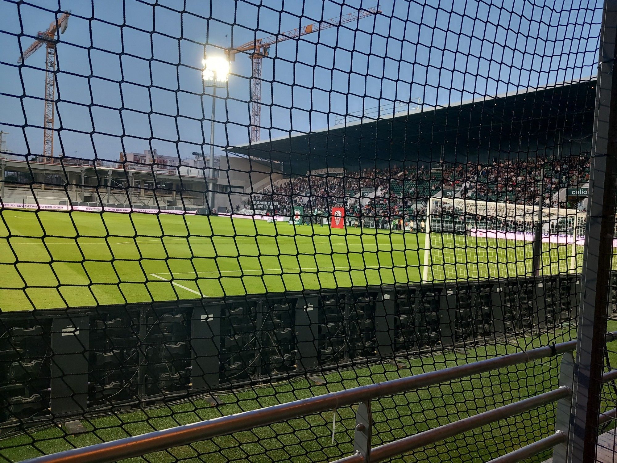 Derrière le filet du parcage, on voit la grande tribune latérale verte. Protocole d'entrée avec les kakémonos logos de clubs. Banderoles contre le diffuseur dans la tribune des ultras.