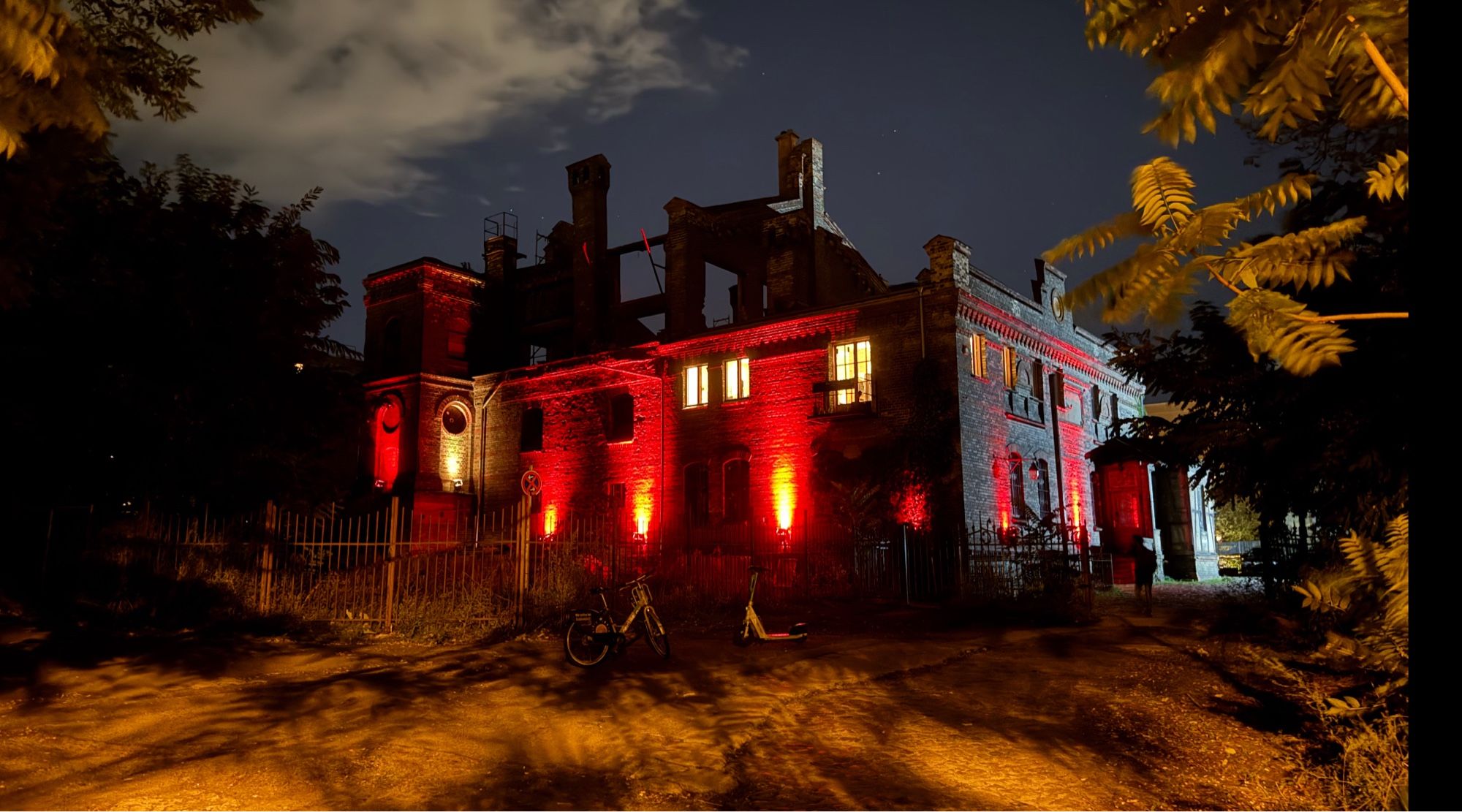 Ruine der alten Brauerei Schweizergarten bei Nacht. Vom Gebäude sind die beiden unteren Etagen erhalten, das Dach infolge von Kriegsschäden nur rudimentär - die Reste heben sich gegen den Himmel ab. Die Fassade ist rot beleuchtet, der Vordergrund durch die Straßenbeleuchtung in Orange. Rechts und links des Gebäudes viele Pflanzen.