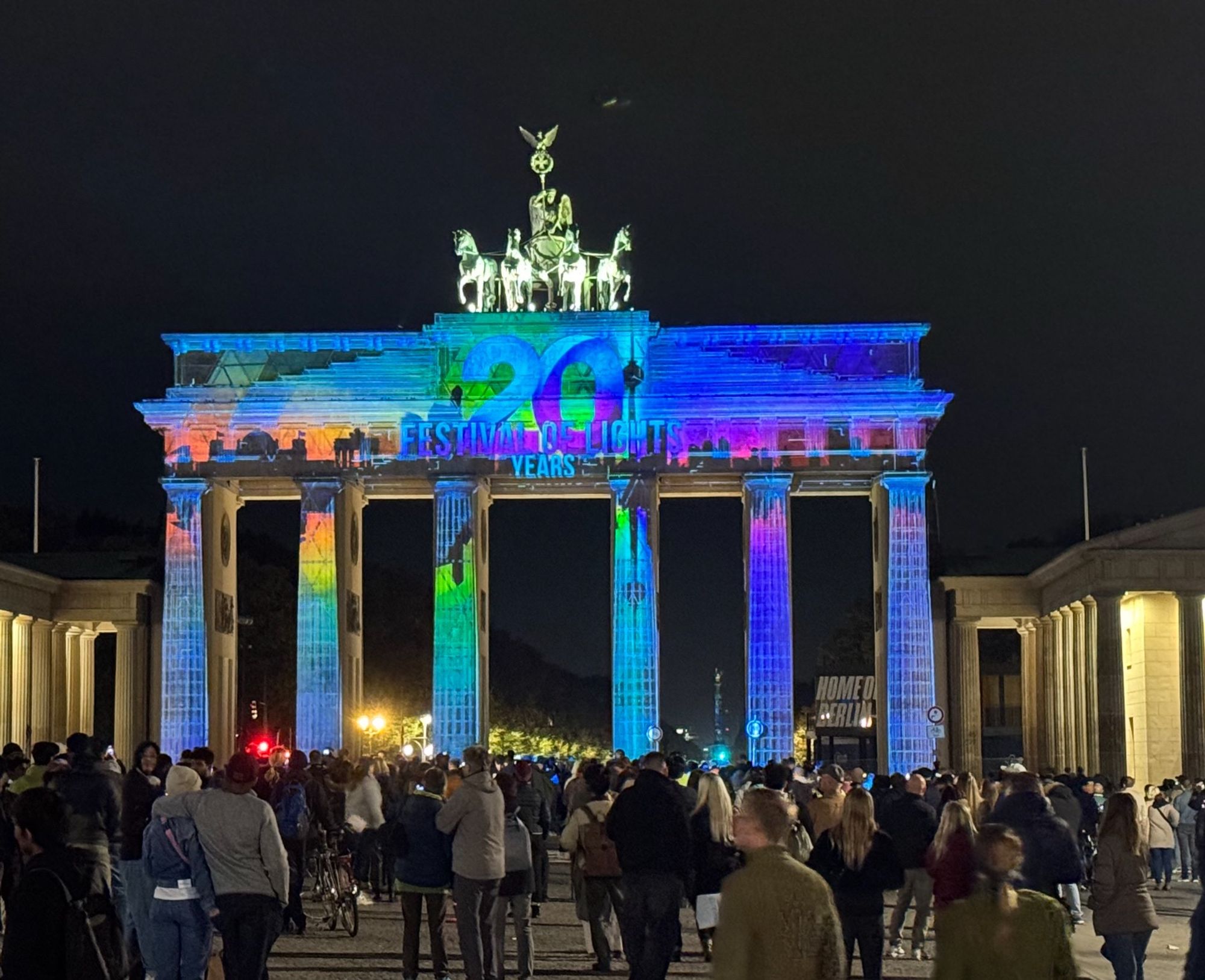 Brandenburger Tor vom Pariser Platz aus vor dunklem Himmel. Auf das eigentliche Tor sind bunte Farben und der Schriftzug "Festival of Lights - 20 Years" projiziert.