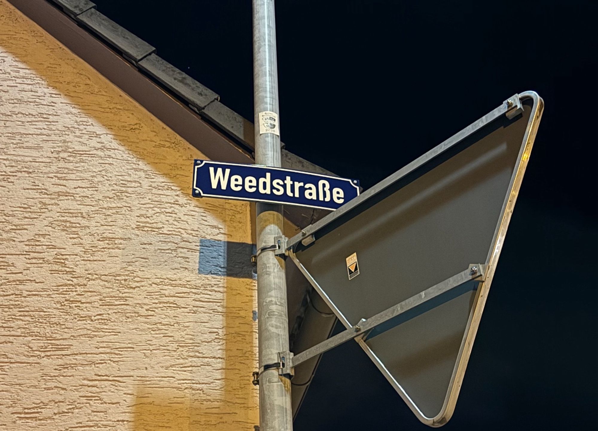 Straßenschild mit der Aufschrift "Weedstraße", vor dunklem Himmel und einem gelben Haus. Direkt darunter ist ein "Vorfahrt gewähren"-Schild von hinten zu erkennen.