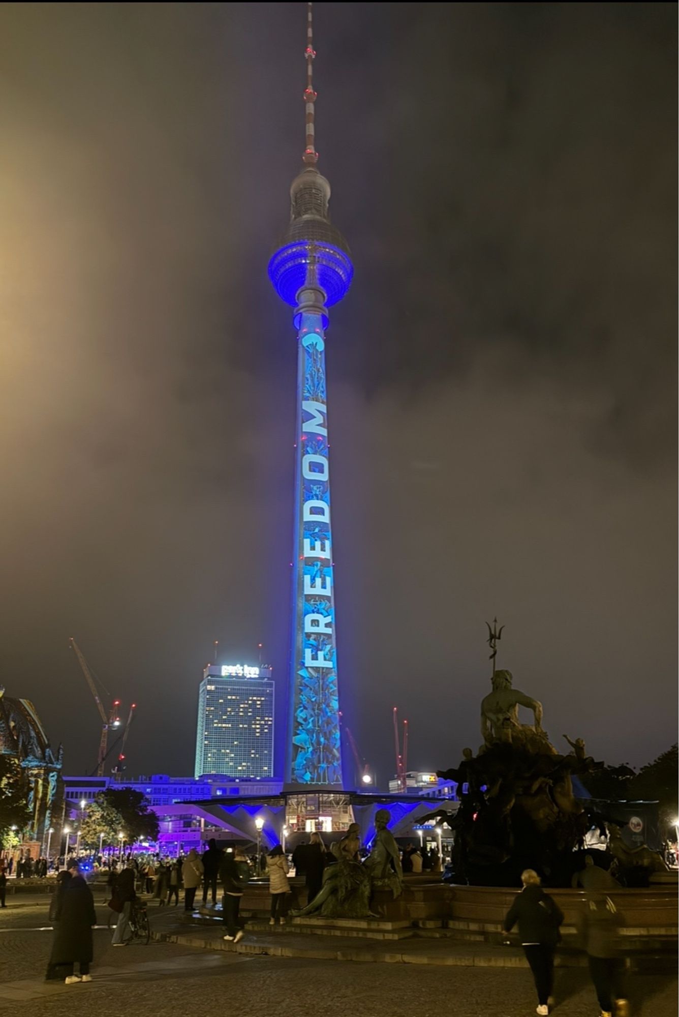 Berliner Fernsehturm von der Westseite vor bewölktem nächtlichen Himmel. Die Kugel ist blau angestrahlt, auf den Schaft ist senkrecht von unten nach oben das Wort "Freedom" projiziert.