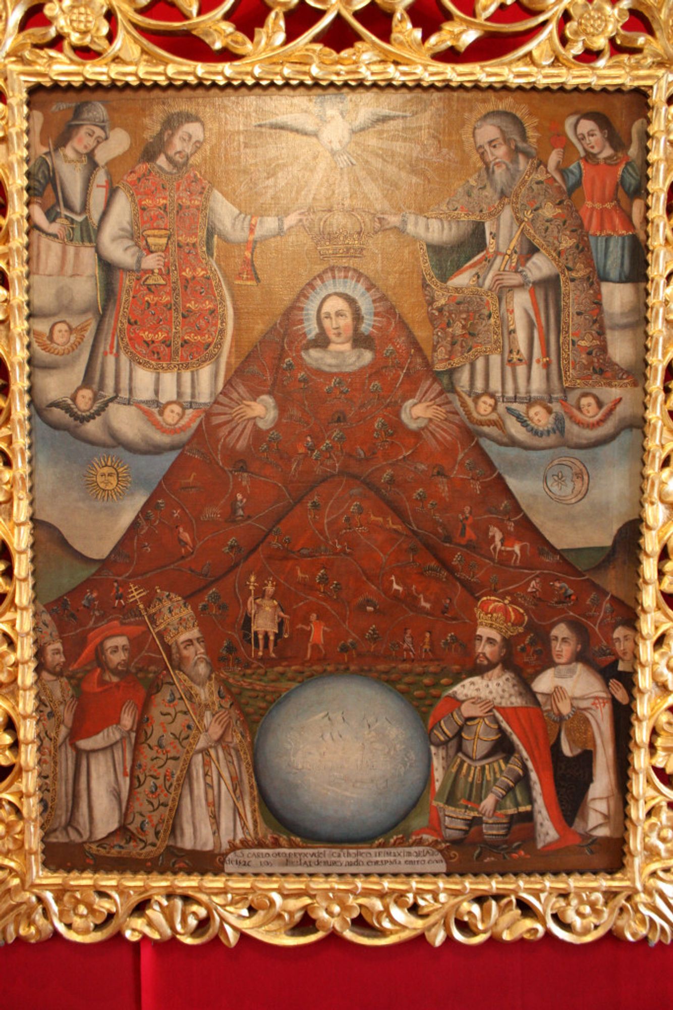 A lavish Baroque religious painting in an ornate gold frame. At the center is a blood red mountain - this forms the "body" of the Virgin Mary, although you can only see her face and her hands protruding from the mountain. Around her men kneeling - seemingly saints, kings, angels, and religious figures such as bishops. A sun and a moon are on either side of the mountain / Mary. A blue empty-looking globe is at the bottom of the painting and a dove is at the top.