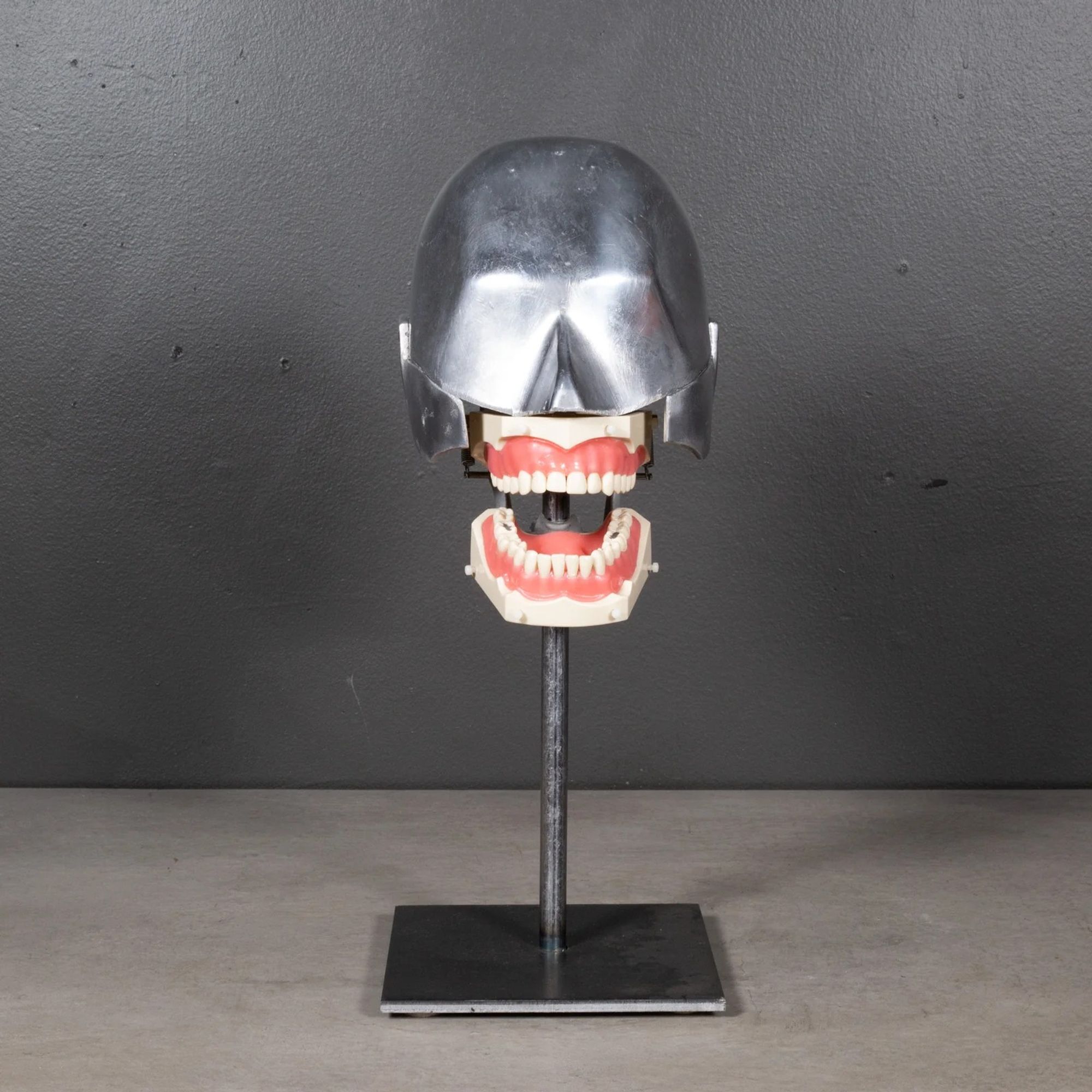 Dental phantom. A head (half silver metal from the nose up; half dentures) on a metal rod and base. There are some fillings visible in the white teeth in the pink gums of the dentures. The nose, ears, and eyebrows are gestured at but not delineated in the metal.