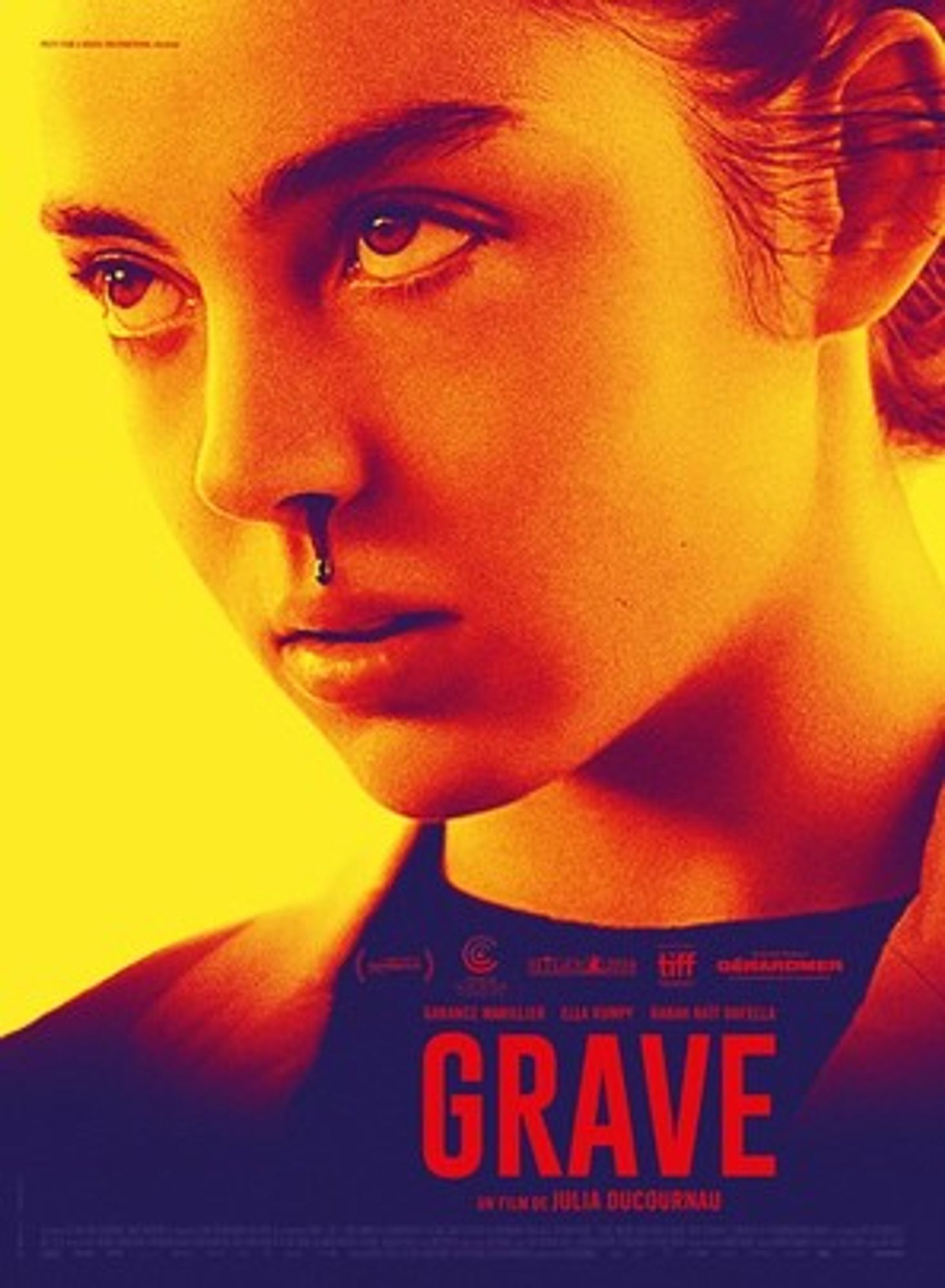 Movie poster for 'Grave,' with the film title in red at the bottom of the poster. With a yellow background, the focus is on a young girl's face. She looks intensely upwards, with her face tilted downwards, with her left nostril dripping blood.