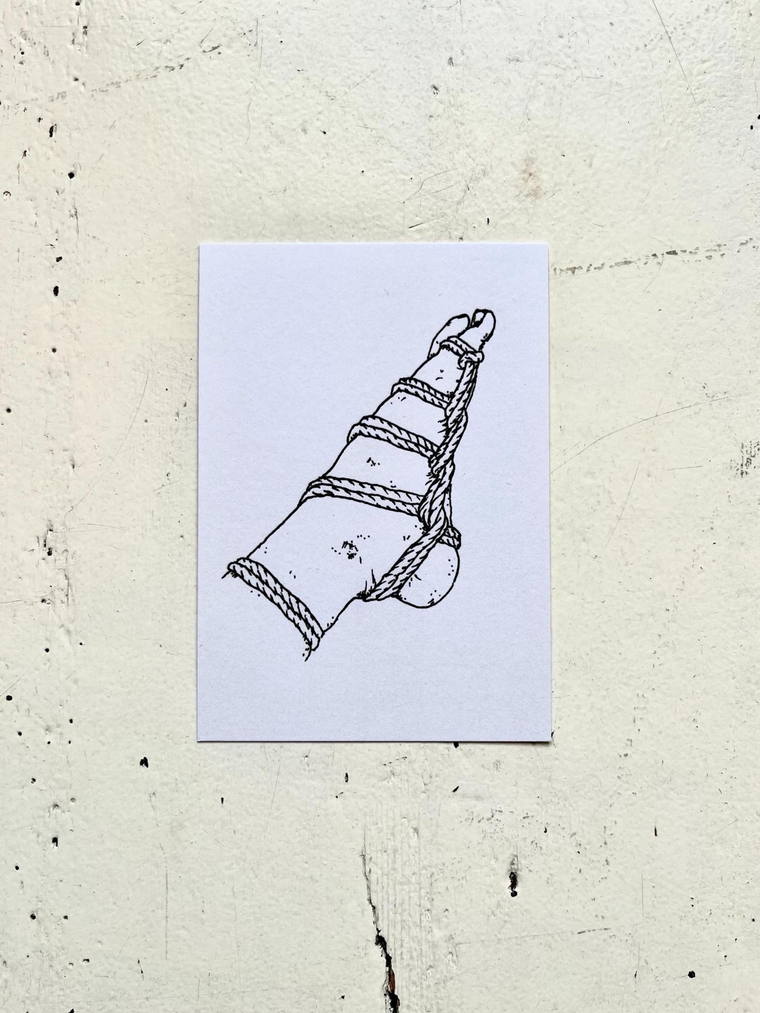 Picture of a postcard on a white wooden background. The postcard shows a black ink drawing on a white background. The drawing features a bondaged foot. The rope holds it in a pointed position like it’s dancing.