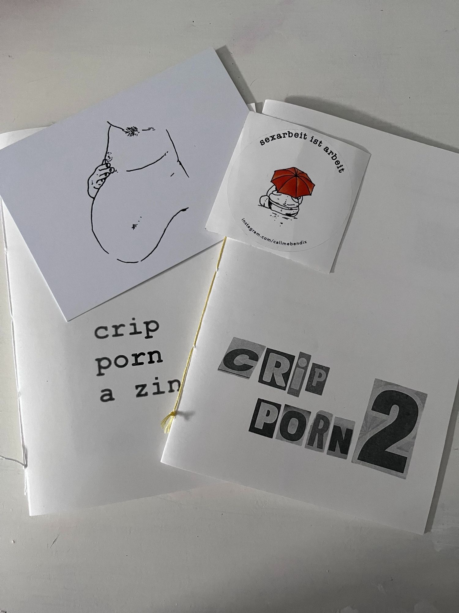 Foto von meiner Kunst. Auf einem kleinen Haufen liegen meine Zines Crip Porn 1 und 2, eine Postkarte von einem dicken Bauch und ein Sticker mit rotem Regenschirm und dem Satz Sexarbeit ist Arbeit.