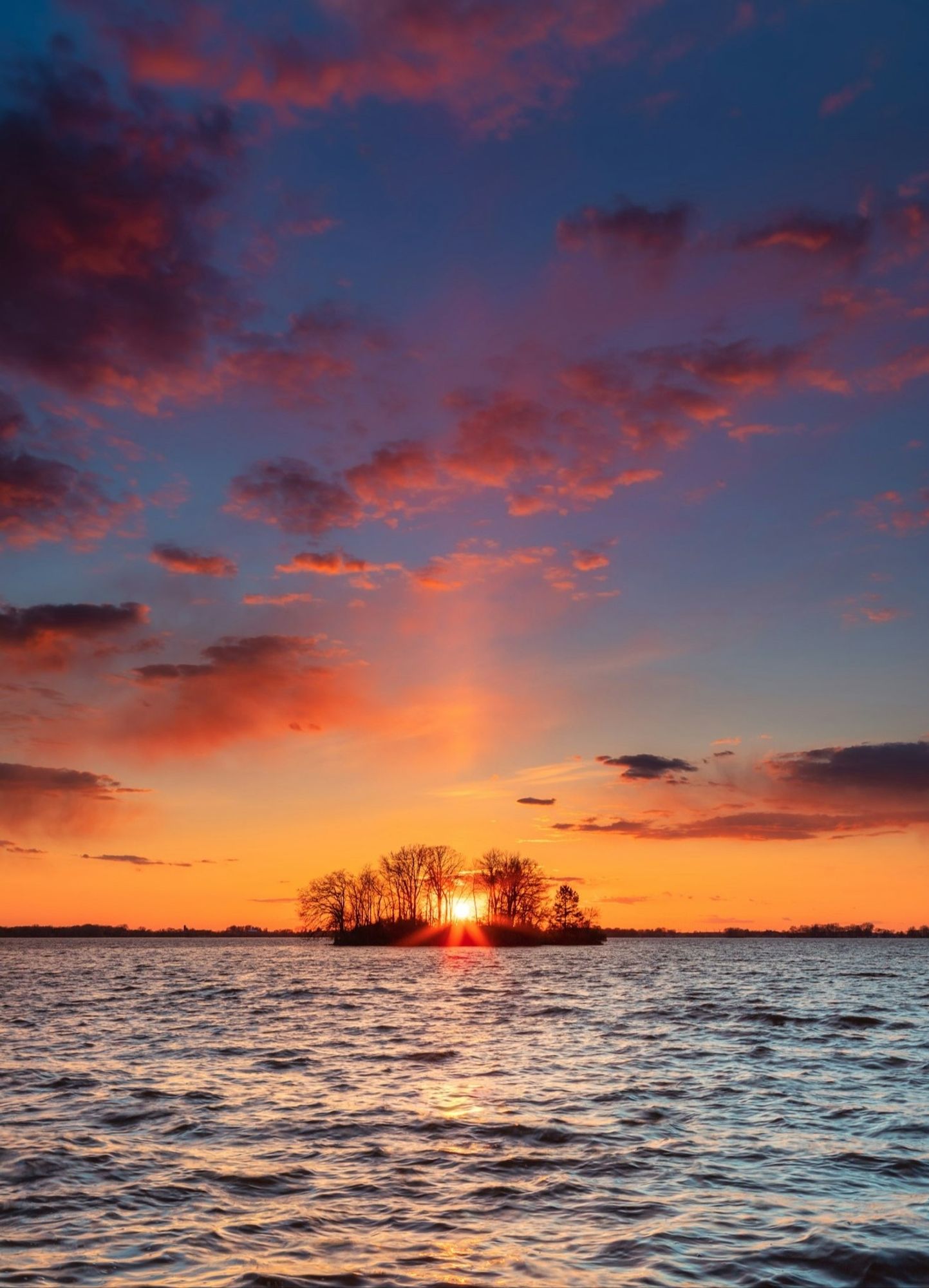 Water in front, at the bottom some trees and the sun that appears behind, coloring the clouds in orange in a blue sky.
📸: Dave Hoefler.