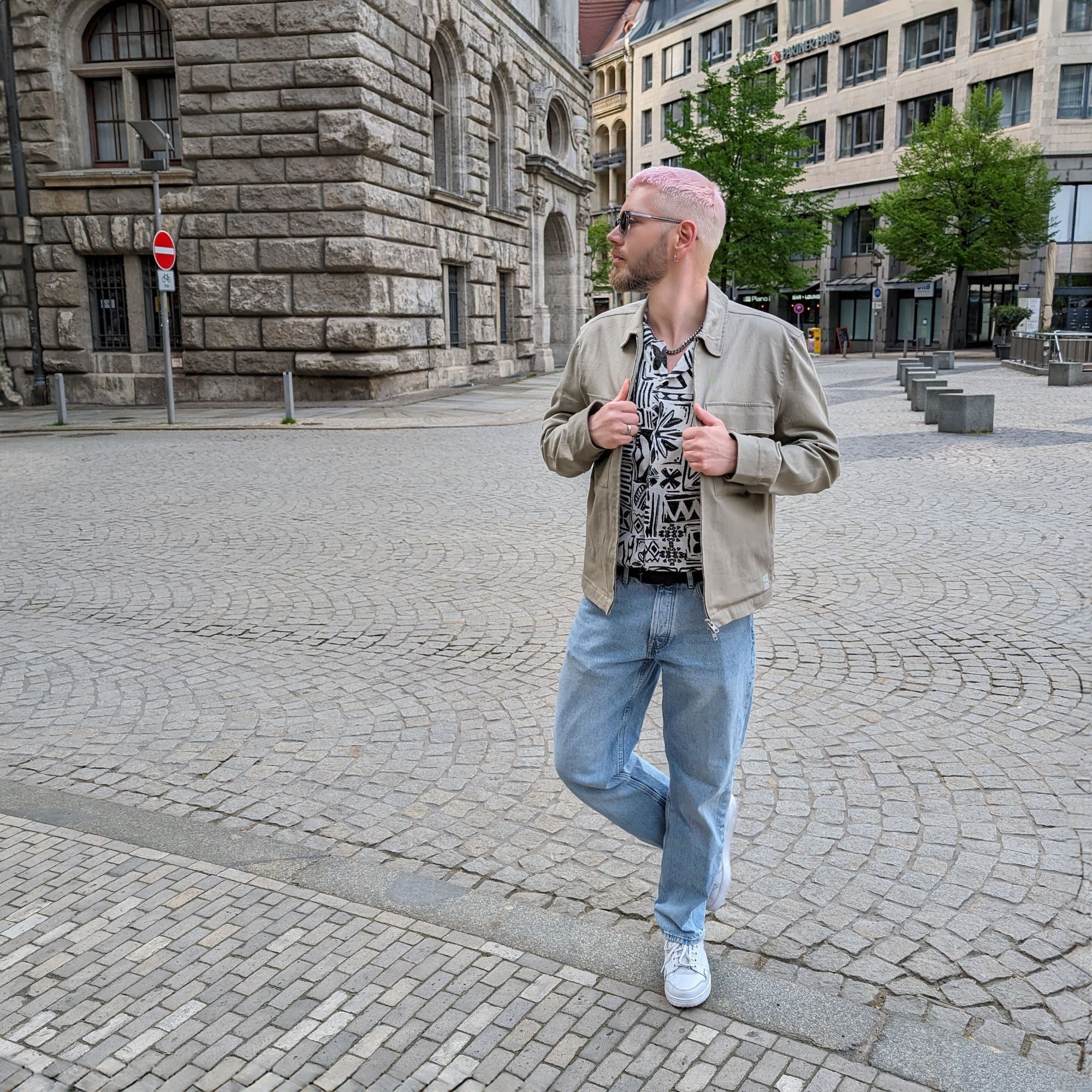 Ich steh in Modelpose vor einem Stadtpanorma mit einer blue Jeans, einsm Hemd mit Muster und einer grauen Jacke. Hände an der Jacke und zur Seite schauend.