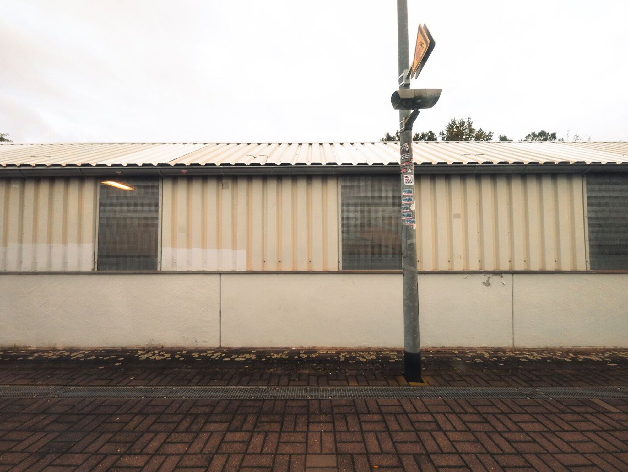 Image Description:
A stark, industrial scene. A long, rectangular building with a corrugated metal roof and white painted walls dominates the frame. The building has several evenly spaced windows with slatted blinds. A solitary lamppost stands to the right, adorned with a street sign and a security camera. The ground is paved with reddish-brown bricks. The overall mood is somewhat bleak and impersonal.