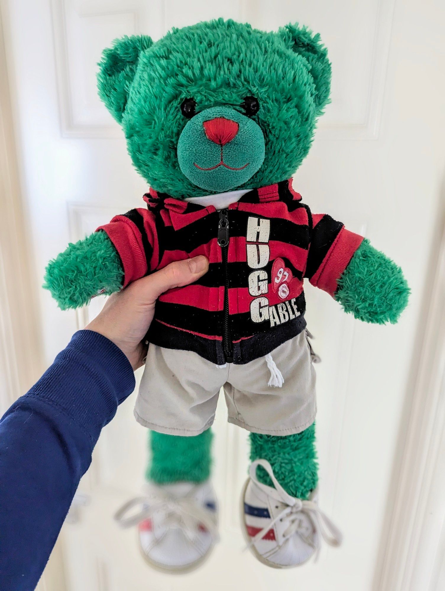 I'm holding a BABW bear in my hand, he is well loved and in a vibrant green colour with a red nose. The bear is wearing a black and red striped hoodie which says "HUGGABLE 97", cream shorts and sneakers.