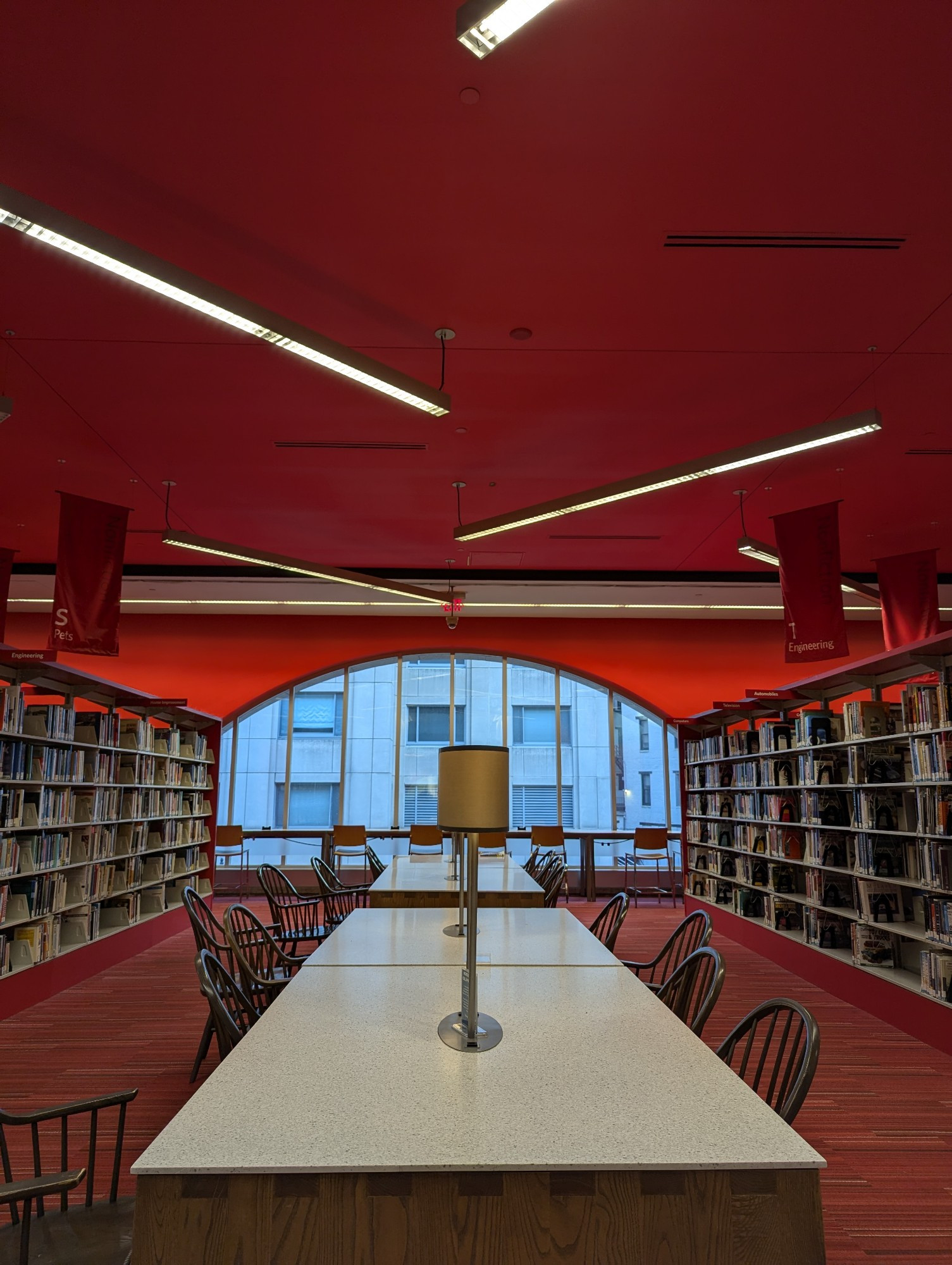 Second floor of the library right as they kicked us off. The ceiling and carpet are red. Bookshelves frame the left and right. In the center are large tables with wooden slatted chairs. In the background is a large arched window with a high top table and chairs.