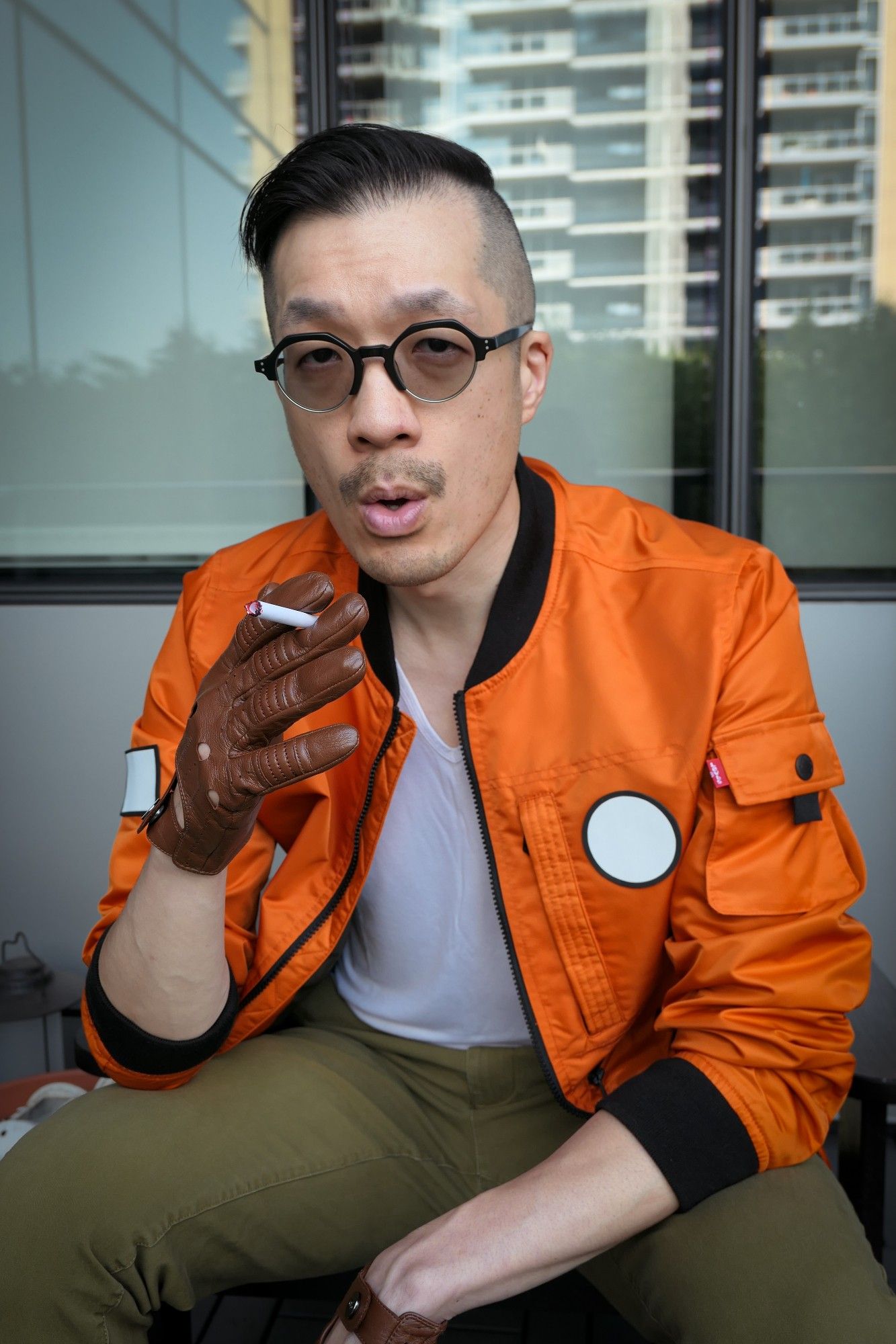 Photo of me dressed as Kim Kitsuragi from Disco Elysium. I'm a middle-aged Asian guy in an orange bomber jacket, olive pants, round glasses, and brown leather driving gloves. Sitting shot. I'm looking directly at the camera and pretending to exhale smoke from the fake cigarette I'm holding.