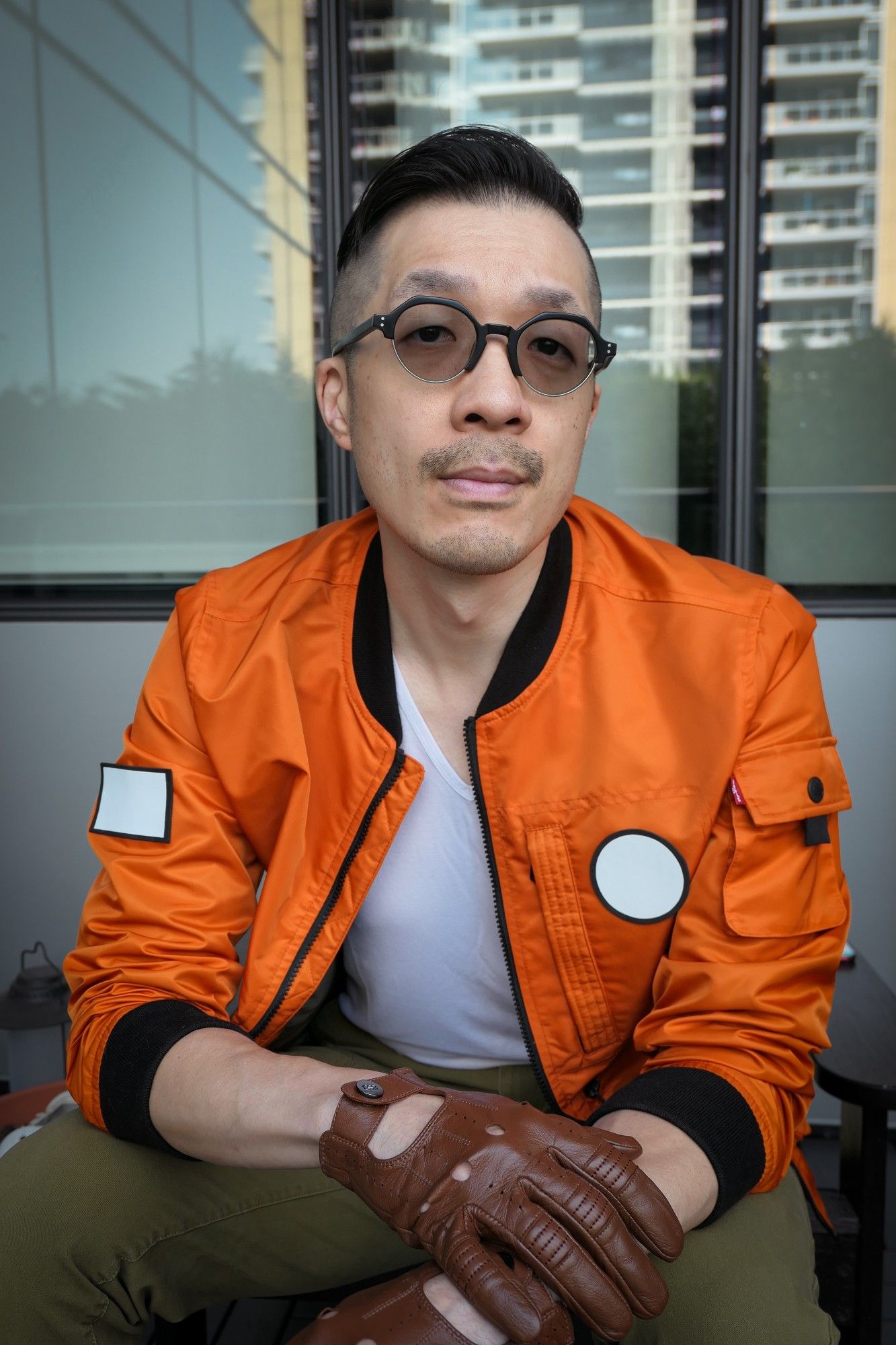 Photo of me dressed as Kim Kitsuragi from Disco Elysium. I'm a middle-aged Asian guy in an orange bomber jacket, olive pants, round glasses, and brown leather driving gloves. Sitting shot. I'm looking at the camera with a slight smile.