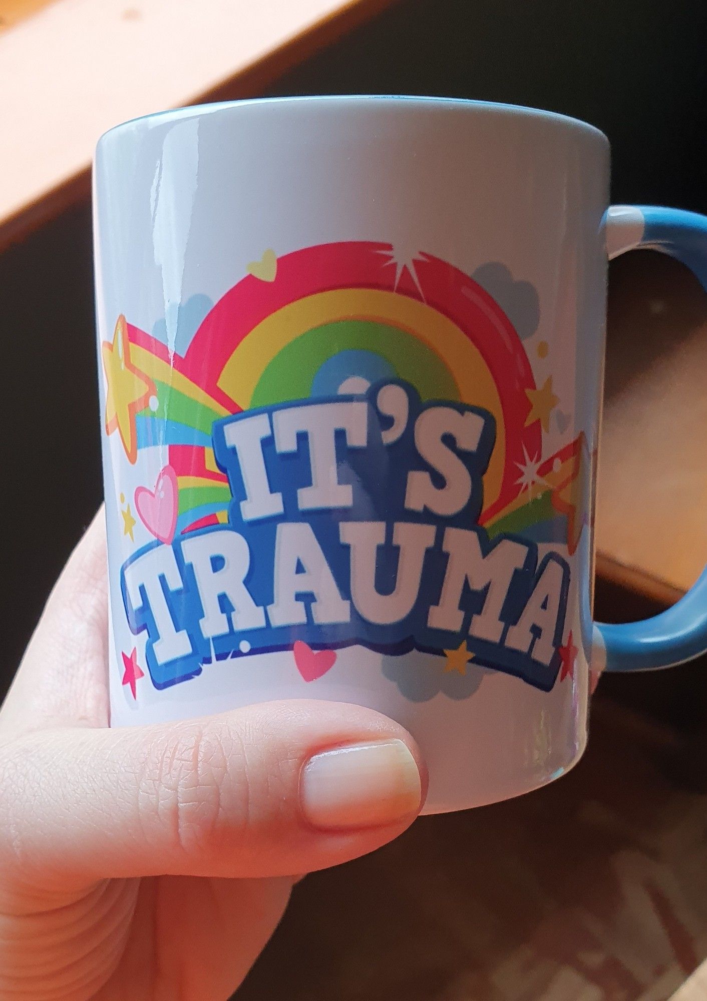 A white woman's hand holding a mug. The mug is white with a large colourful decade that reads "IT'S TRAUMA" surrounded by rainbows, hearts and stars. It's hideous. It's from a Silent Hill game.