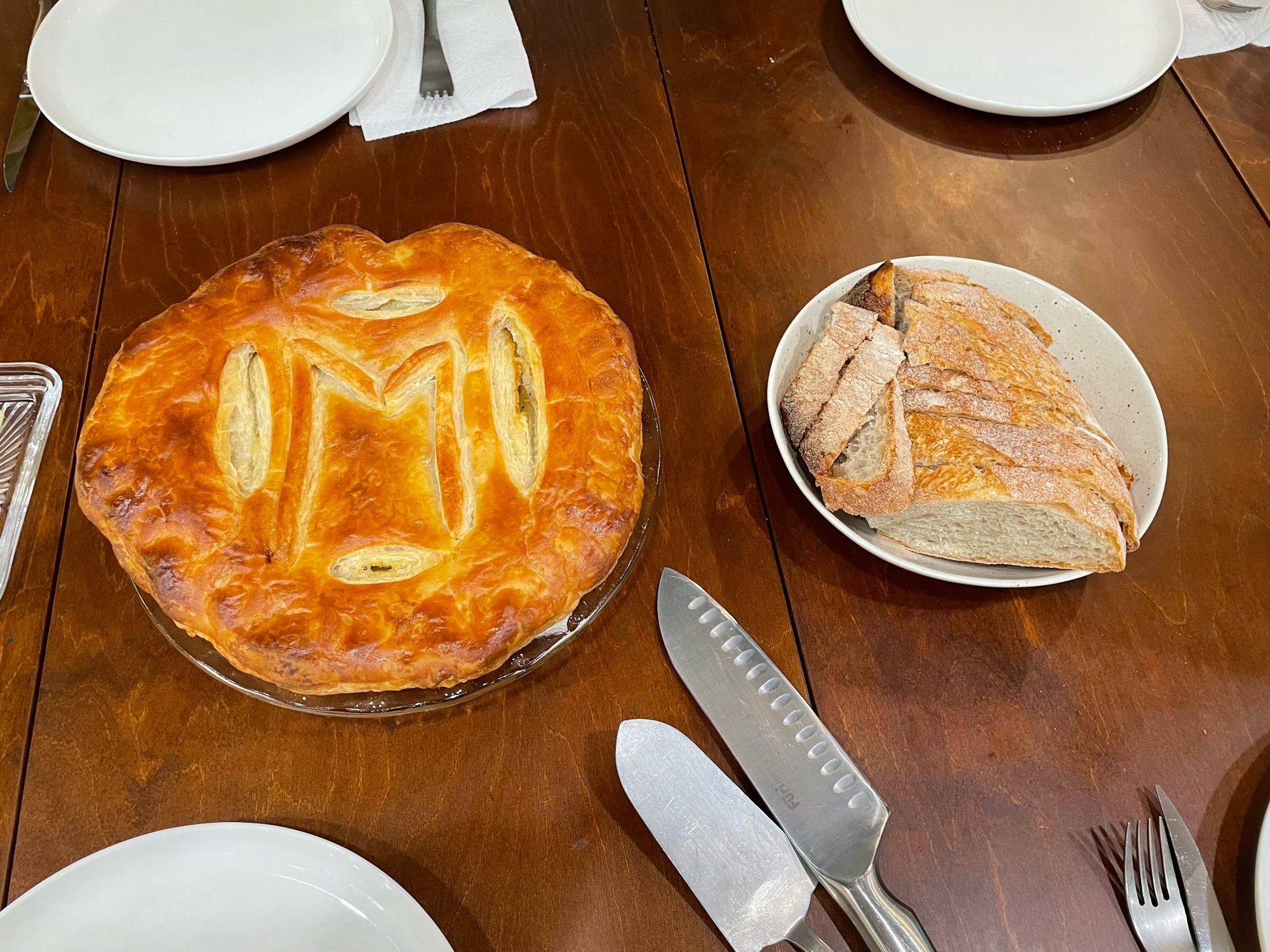 A very balanced meal set on a warm brown wooden table. In the center left is another golden crusted baked chicken pot pie in round glass deep pie dish with a puff pastry M baked into the top and on the right a bowl freshly baked bread slices. The deal is you get what you want for your birthday and this year he wanted carb on carb action