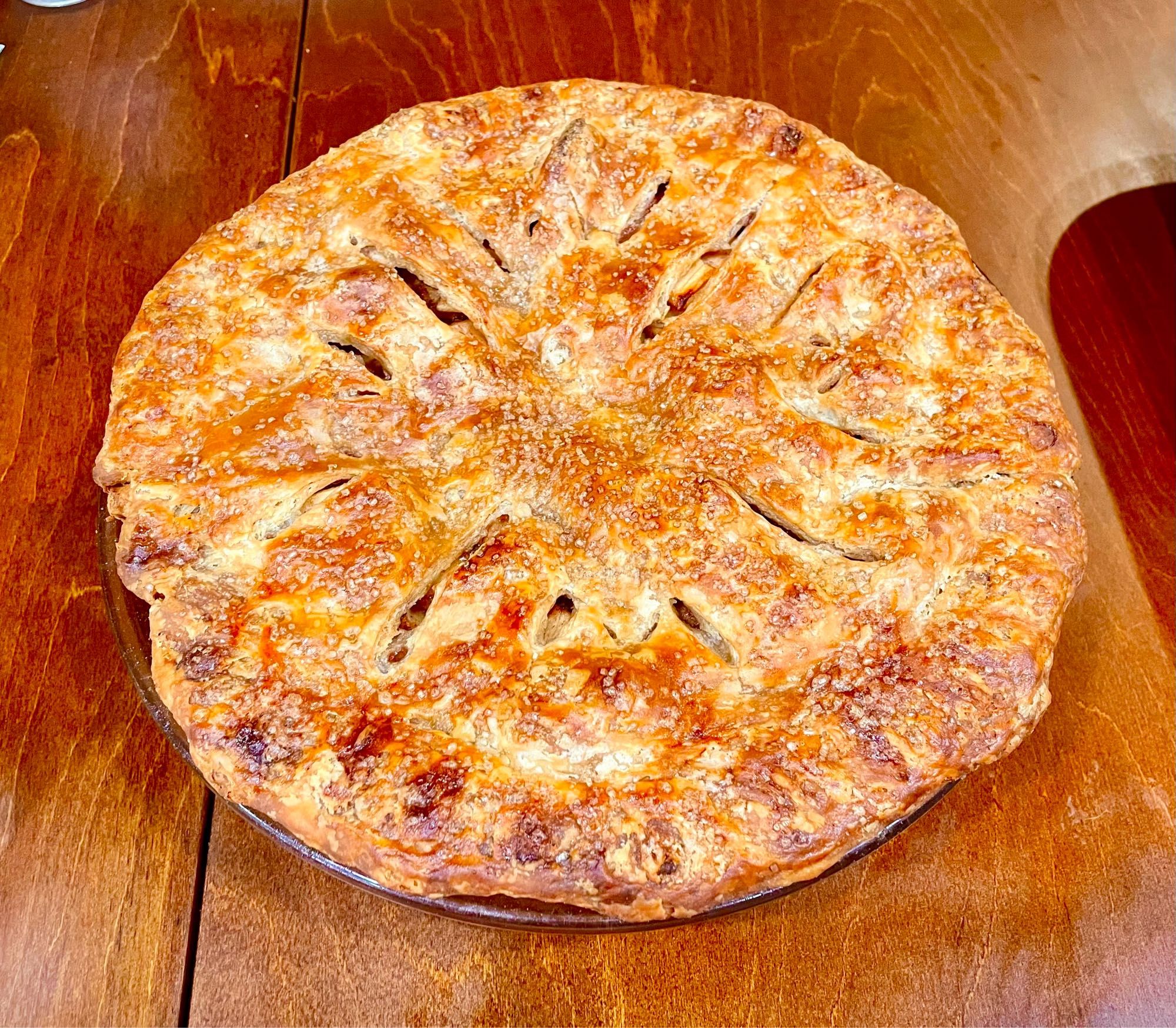 Another golden crusted baked pie in a glass dish. This one is an apple pie with coarse turbinado sugar baked into the crust. The vent holes are cut in a circular pattern with the smaller slashes made into M shapes