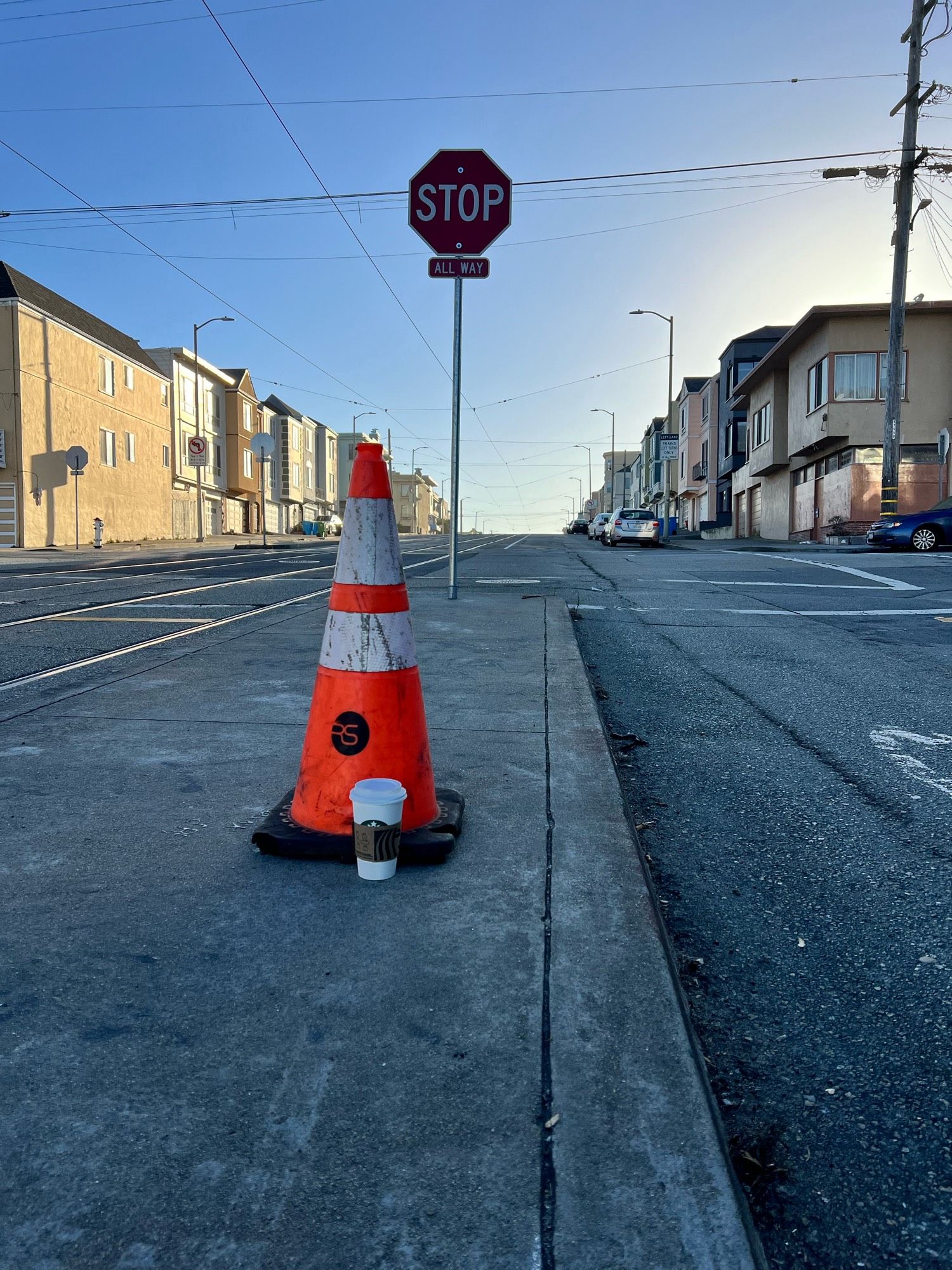 A traffic cone sits on a buss stop curb island on an urban street with the sun at an early angle. There’s a cup of Starbucks just at its base as it it might suddenly lift it up for a sip.