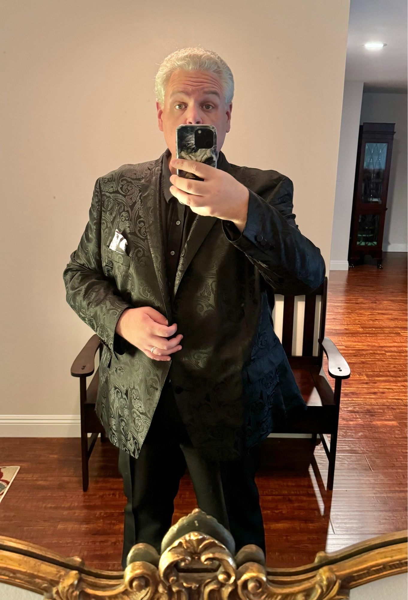 A mirror reflection selfie of a handsome gray and white haired man in a black tuxedo jacket and matching pants and shirt