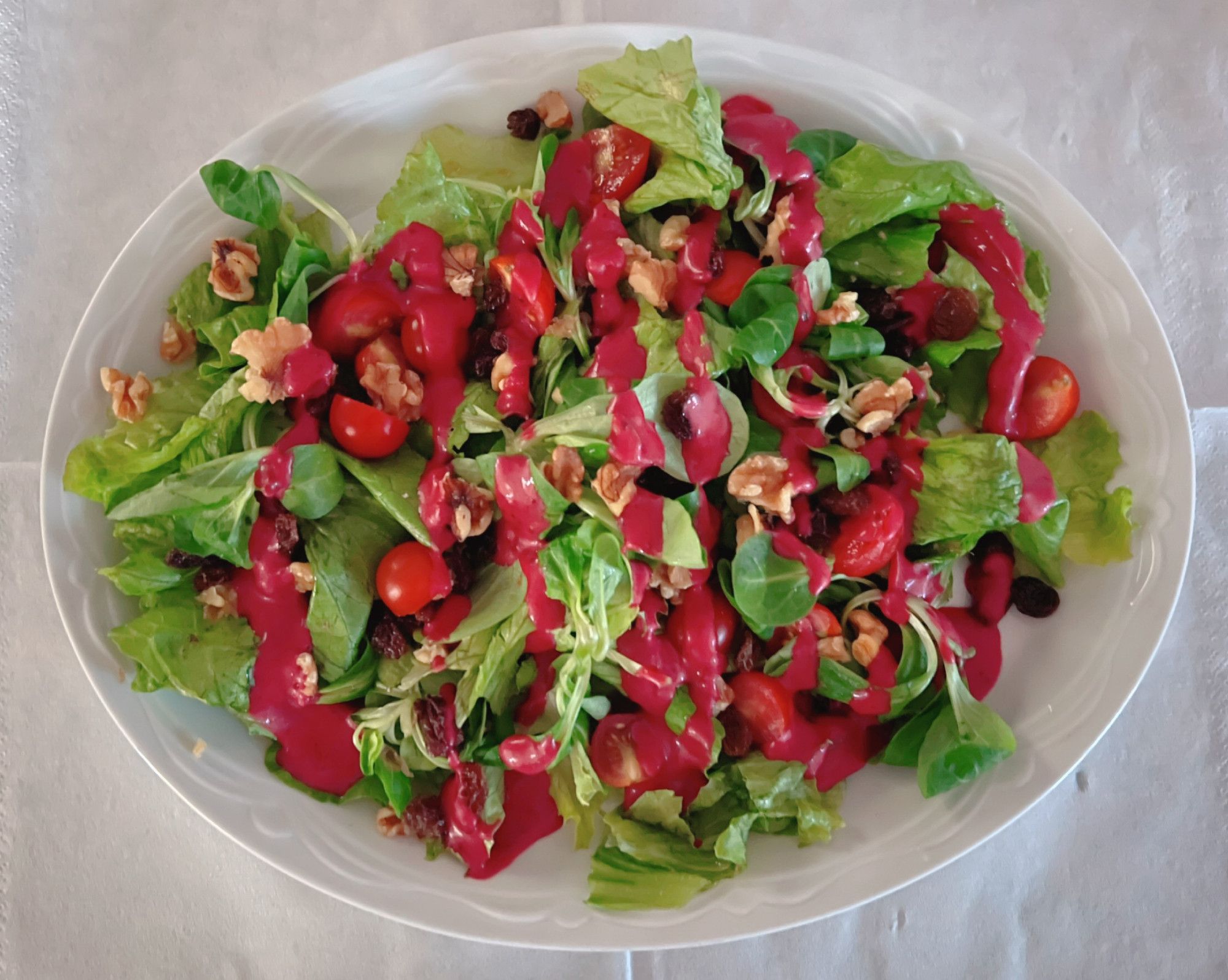 Green salad on a white oval plate. Bed of lettuce and lamb's lettuce with cherry tomatoes, nuts and raisins and a shiny homemade red berries sauce in a zig-zag