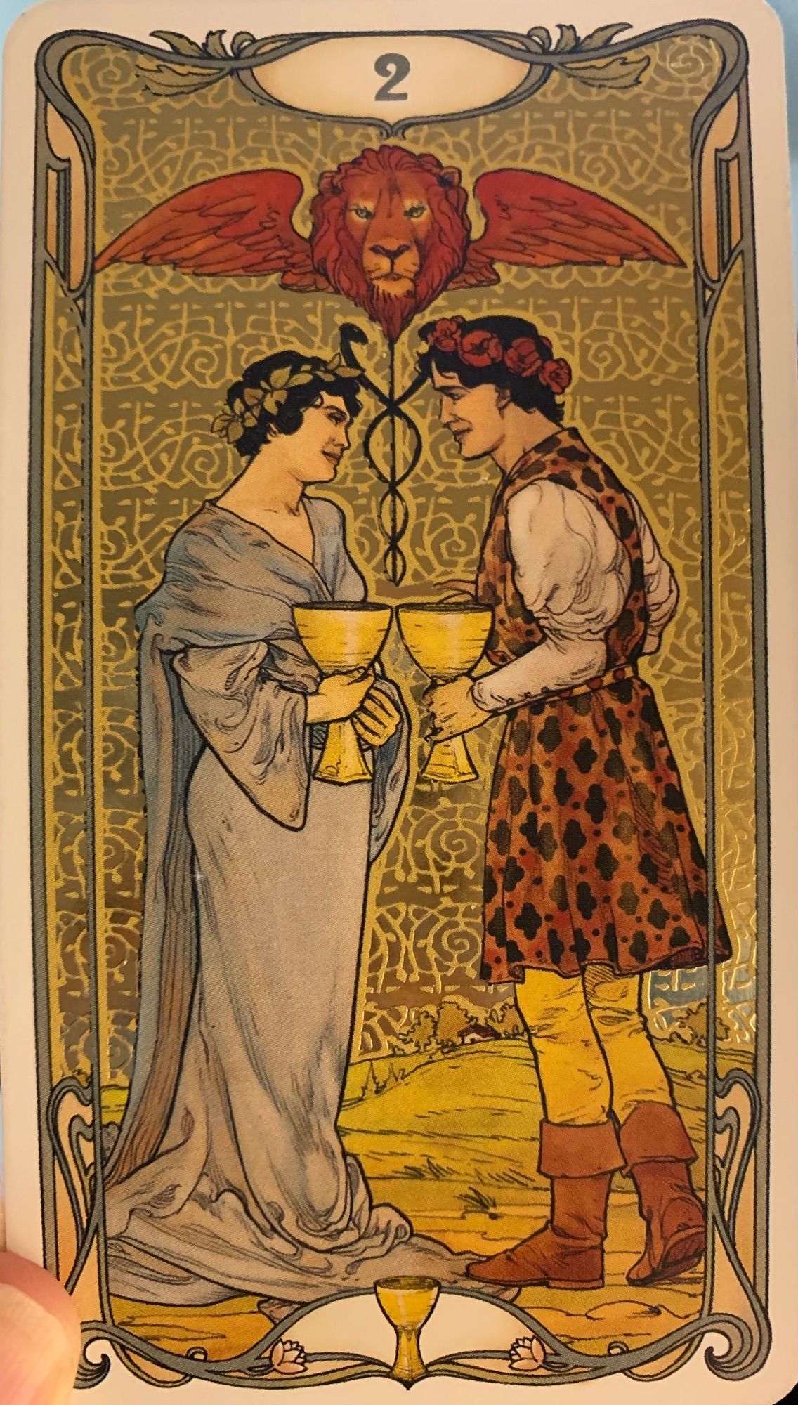 The Two of Cups from the Golden Art Nouveau Tarot deck. Beneath a winged lion's head and a caduceus, a pair of human beings are gazing at each other adoringly. They each hold a golden cup in their hands, and these cups are held together as if for a toast. One is wearing a pale blue gown, the other a patterned tunic over leggings and boots.