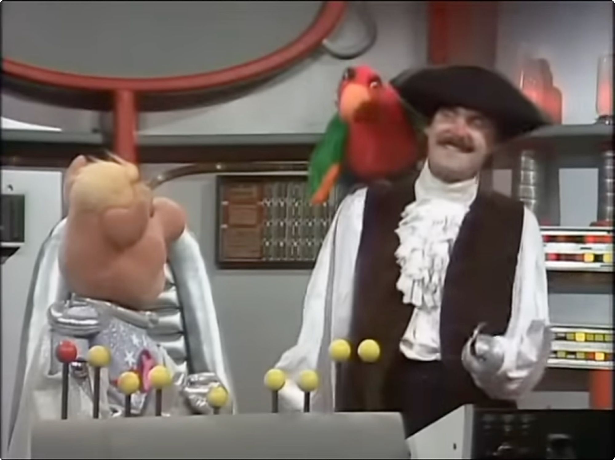 Screen capture of John Cleese, with a tricorn hat, frilly shirt, hook for a hand, and a parrot on his shoulder, confronting Captain Link Hogthrob on the bridge of the space ship Swinetrek, from a Pigs in Space sketch on The Muppet Show.