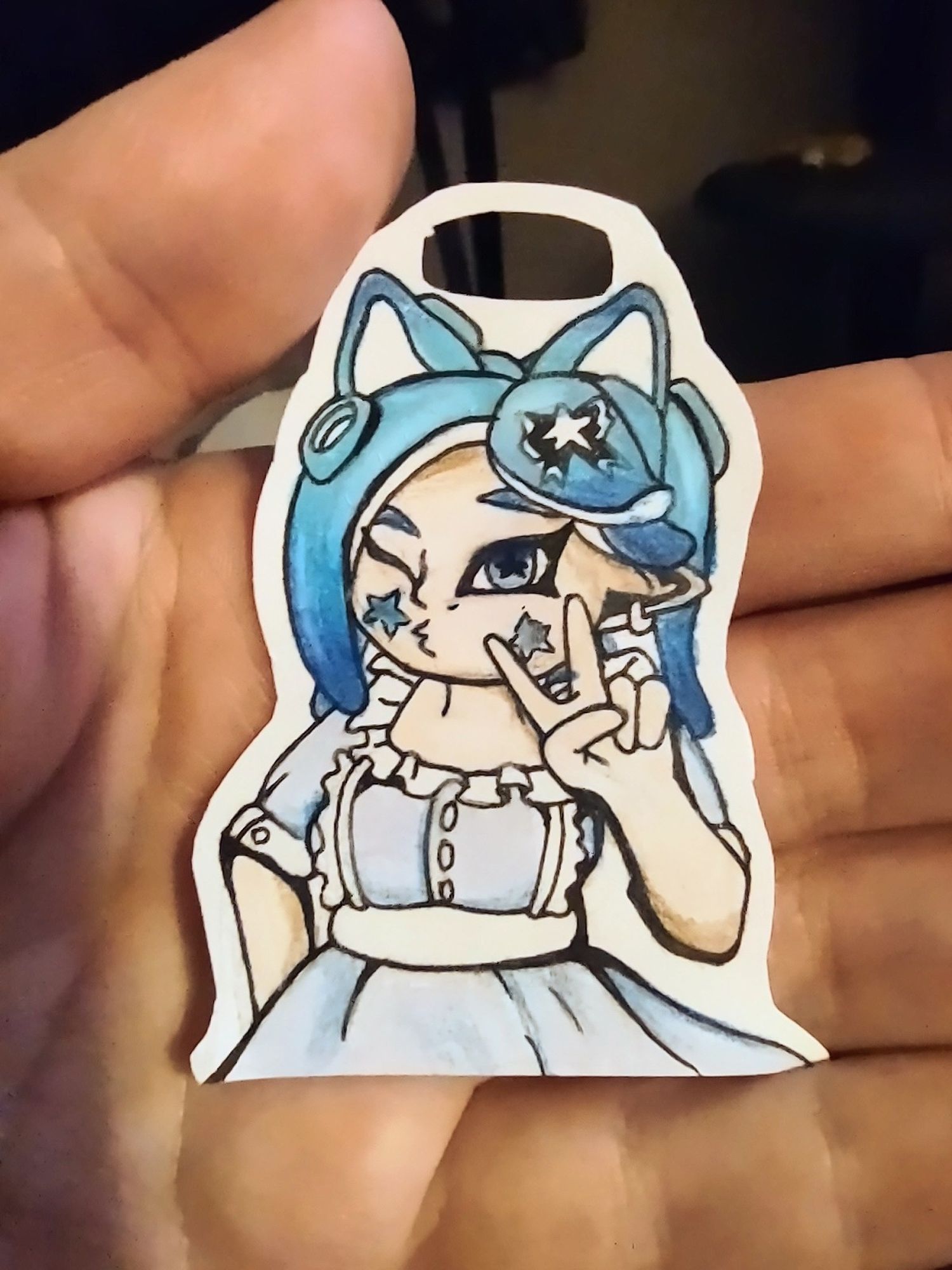 Hand holding a small paper drawing of an original character or fan character from a game called splatoon. She has blue tentacle like hair, pale skin and a blue lolita style dress with white frills.