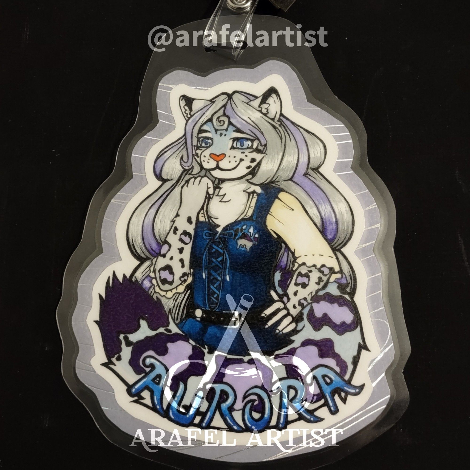 Laminated badge with clip of a purple and blue-grey snow leopard fursona wearing a blue dress. The name Aurora is at the bottom along the tail.