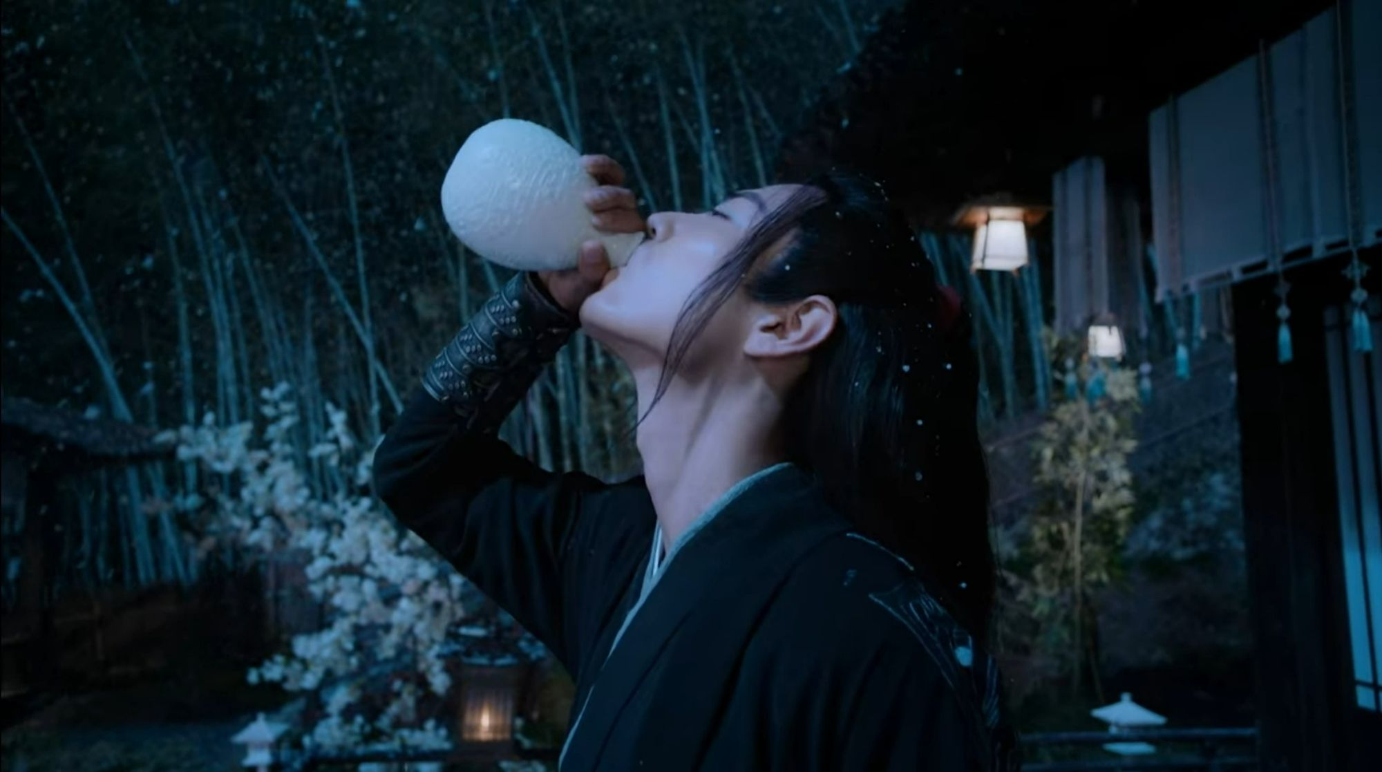 Closeup of Wei Wuxian (actor Xiao Zhan) standing outside while it's snowing, drinking from a bottle. He's in picture from chest up, wearing black robes.