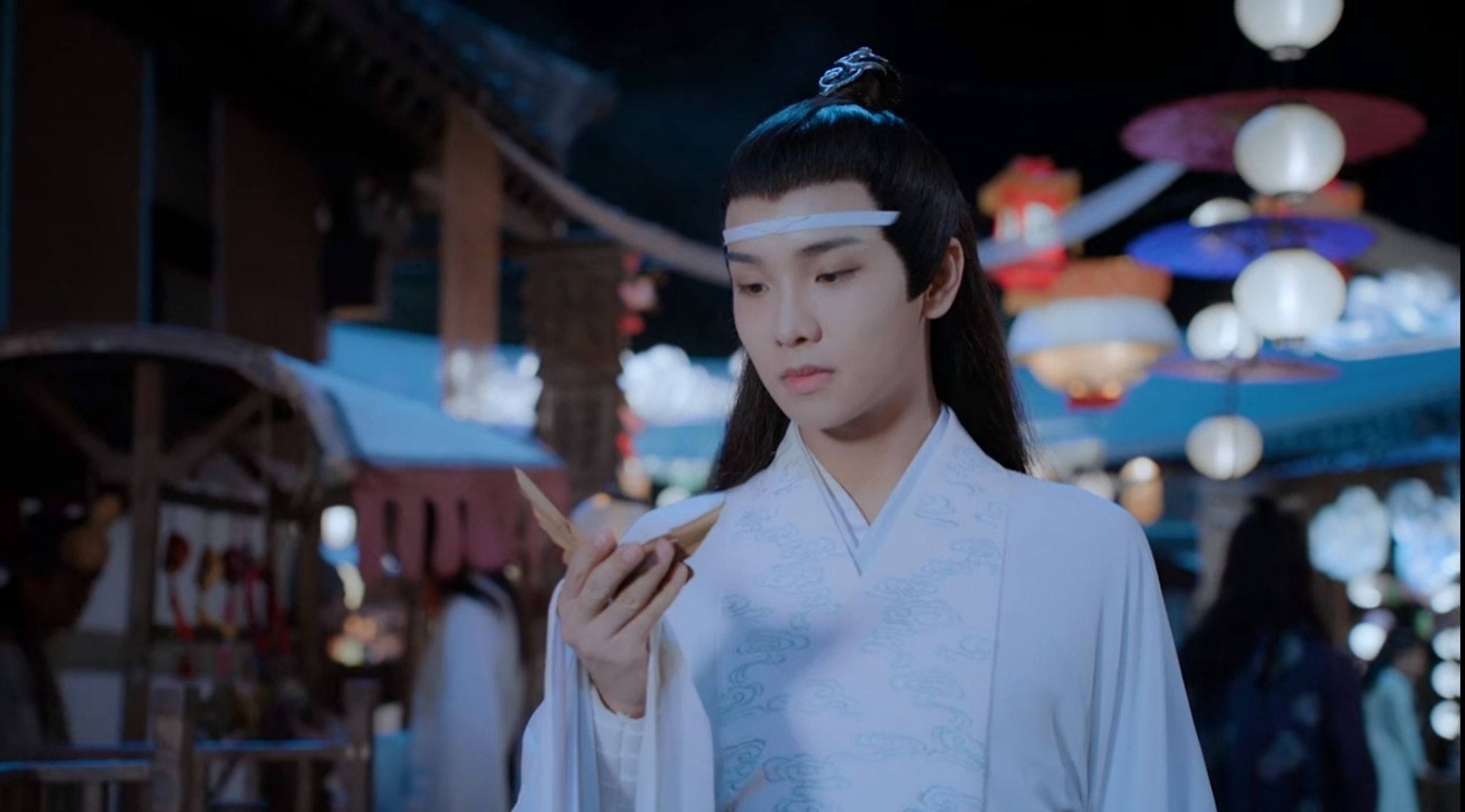 Closeip of Lan Sizhui at a street booth looking at butterfly toy he is holding, lots of lanterns around