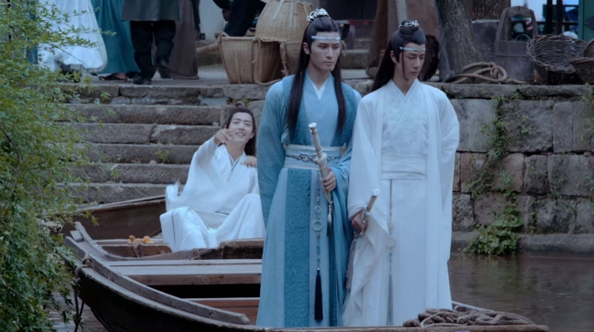 In front Lan Xichen (actor Liu Haikuan) in blue robes and Lan Wangji (actor Wang Yibo) standing on a boat. Behind them, in another boat is Wei Wuxian (actor Xiao Zhan) in white robes throwing (a loquat) towards the Lan brothers.