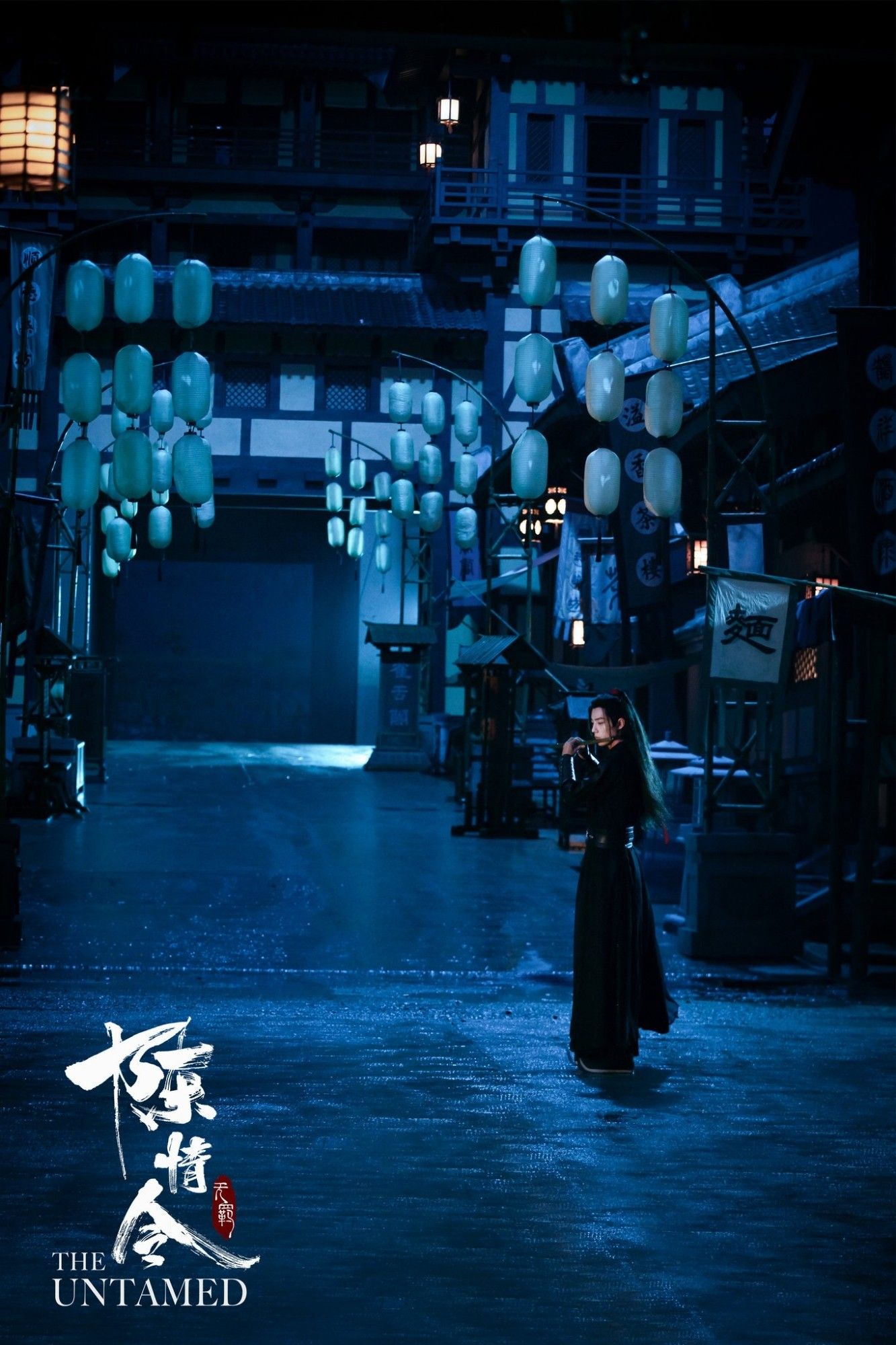 Wei Wuxian (actor Xiao Zhan) standing in middle of steet in empty night time town playing his dizi. He is wearing black robes and the whole picture is blue shade.