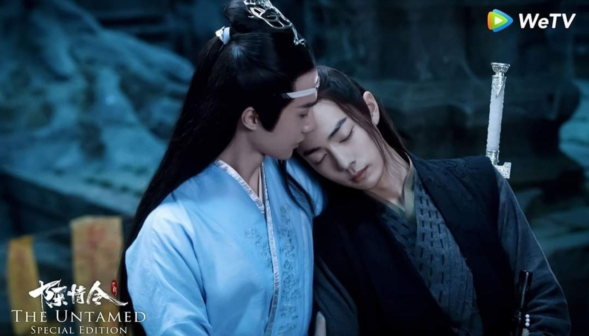 Wei Wuxian (actor Xiao Zhan) leaning his head to Lan Wangji's (actor Wang Yibo) shoulder with his eyes closed. Lan Wangji is looking at Wei Wuxian.  Lan Wangji in light blue robes and Wei Wuxian in black and gray robes. Both in picture from waist up.