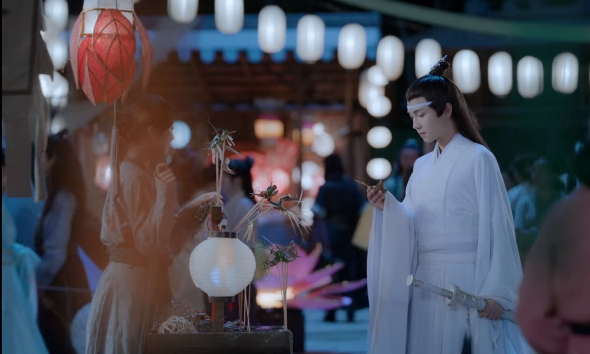 Lan Sizhui at a street booth looking at something he is holding, lots of lanterns around